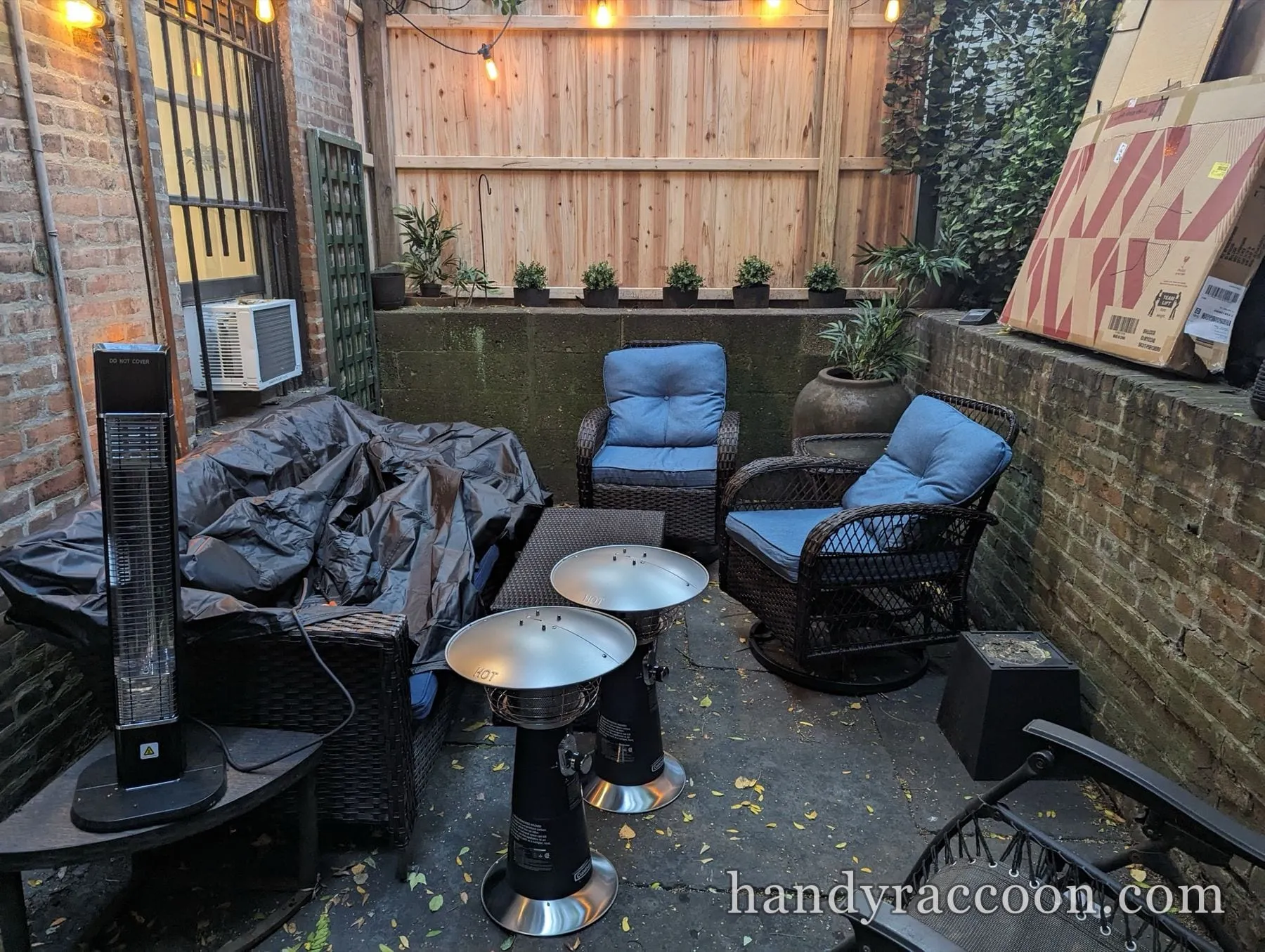 Dusk backyard patio with brown wicker chairs, blue cushions and two tabletop gas heaters under string lights.