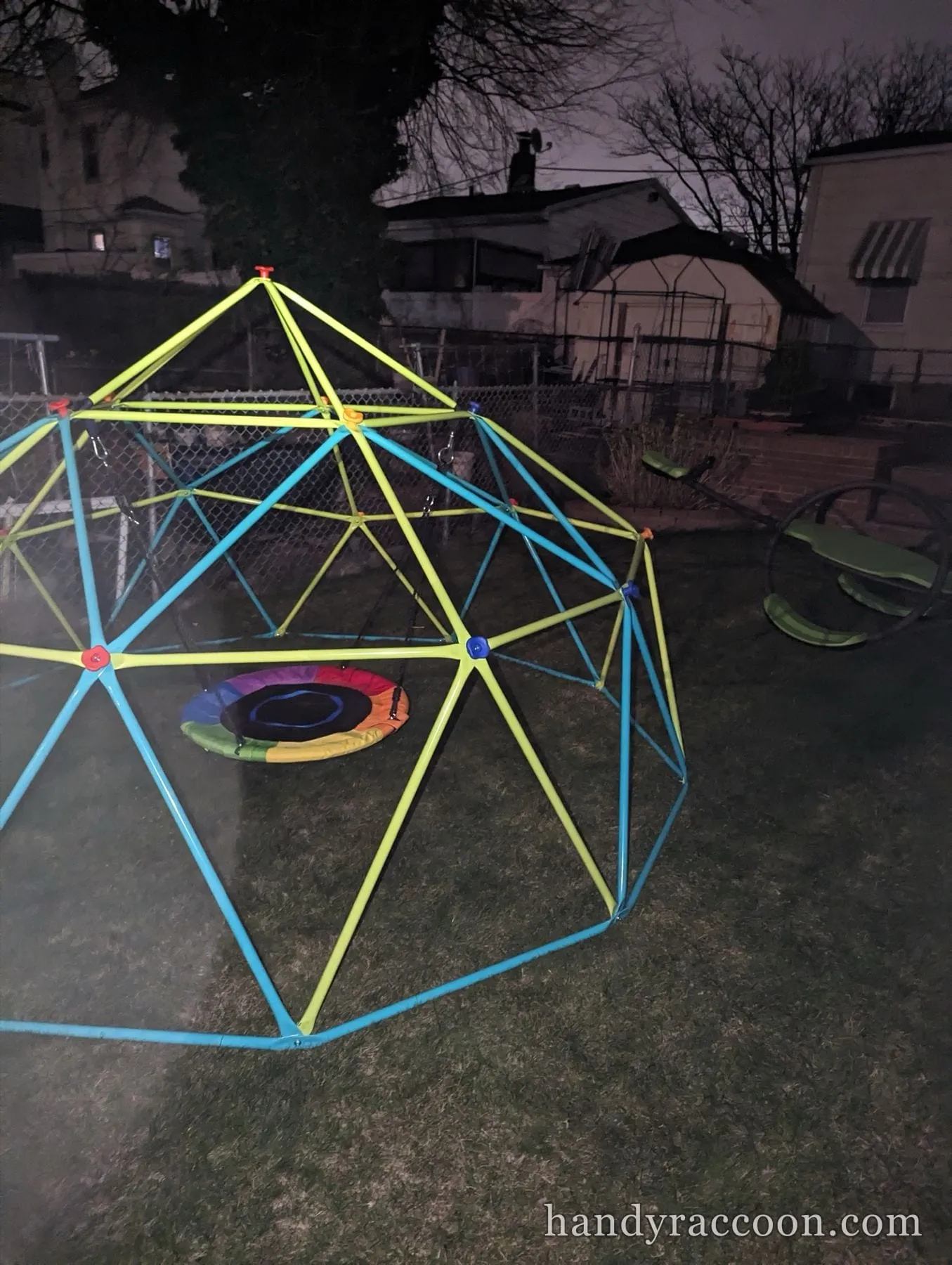 Assembled green and blue geo dome climber with a round nest swing hanging inside, on a lawn at night.