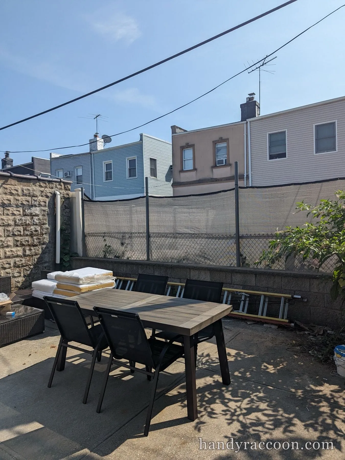Assembled wood-top patio dining table with black sling chairs on a concrete yard, row houses behind.