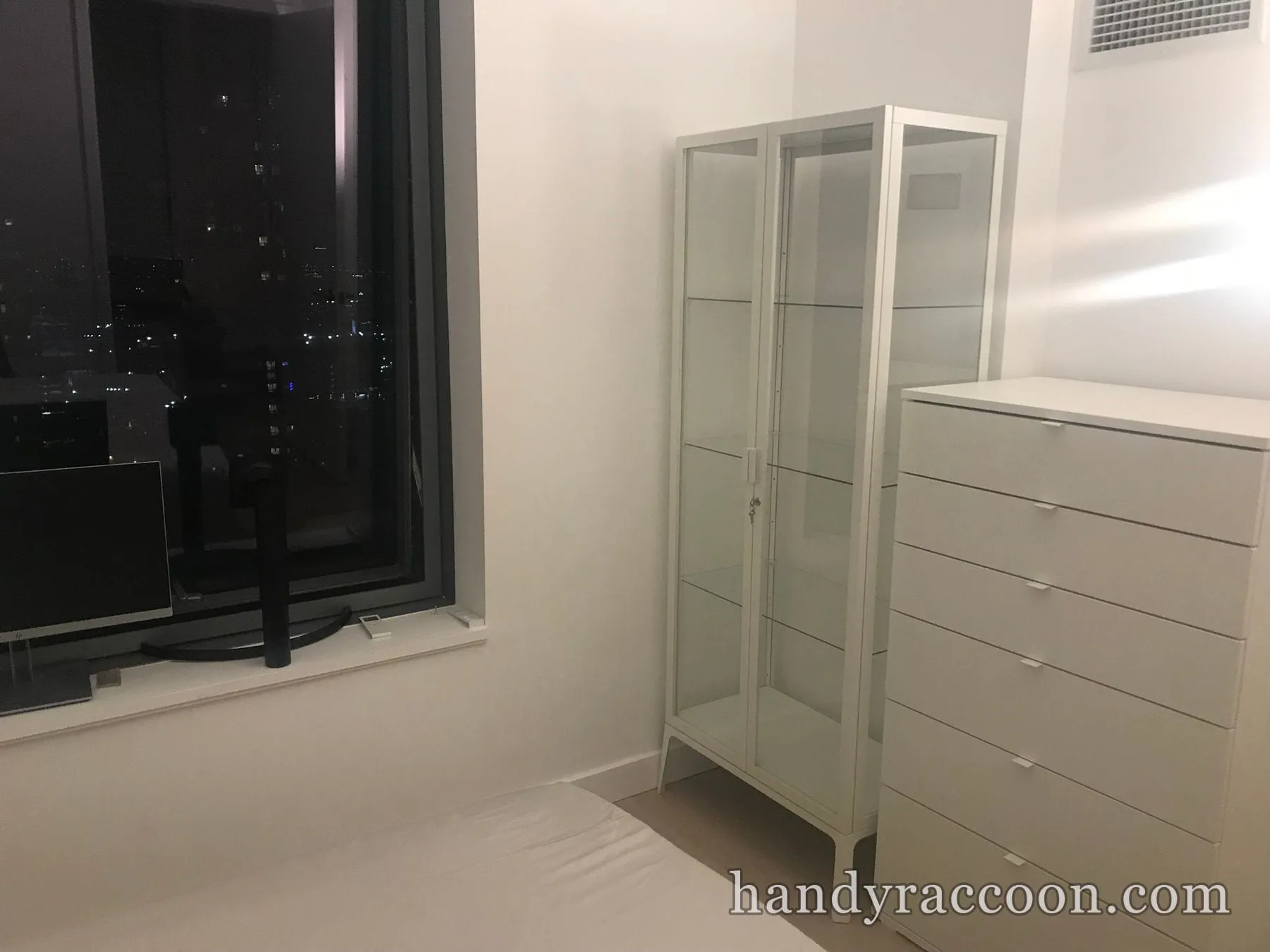 White glass-front display cabinet beside a tall white six-drawer dresser in a bedroom at night