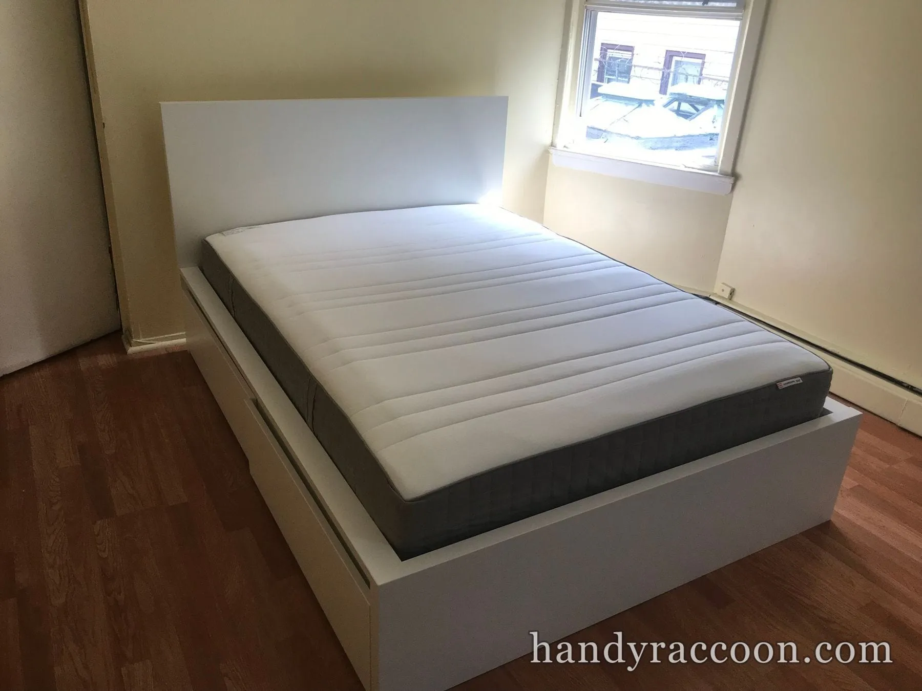 White platform bed with headboard and grey mattress assembled in an empty room by a window