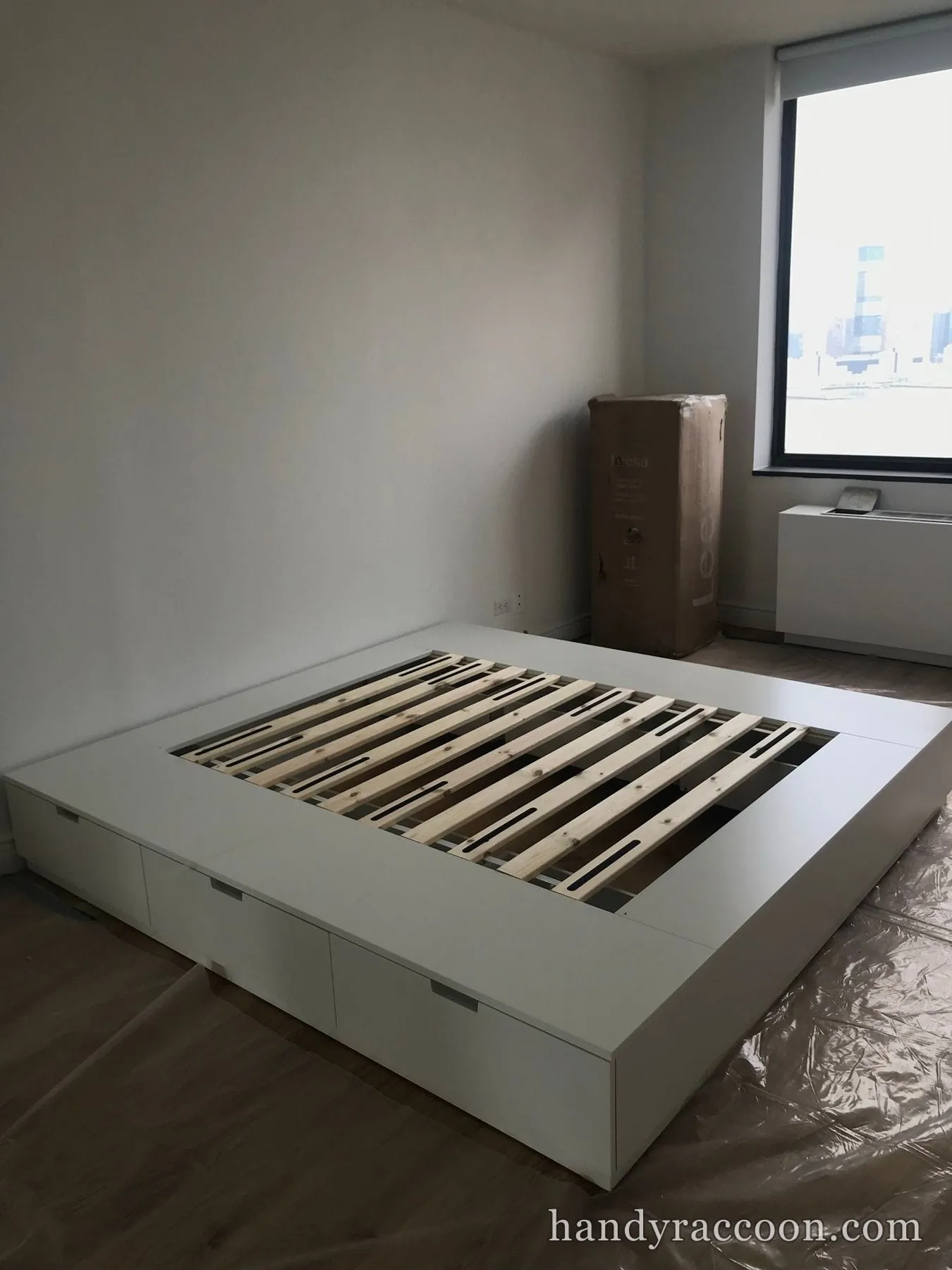 White platform bed base with storage drawers and wood slats being installed in an empty room