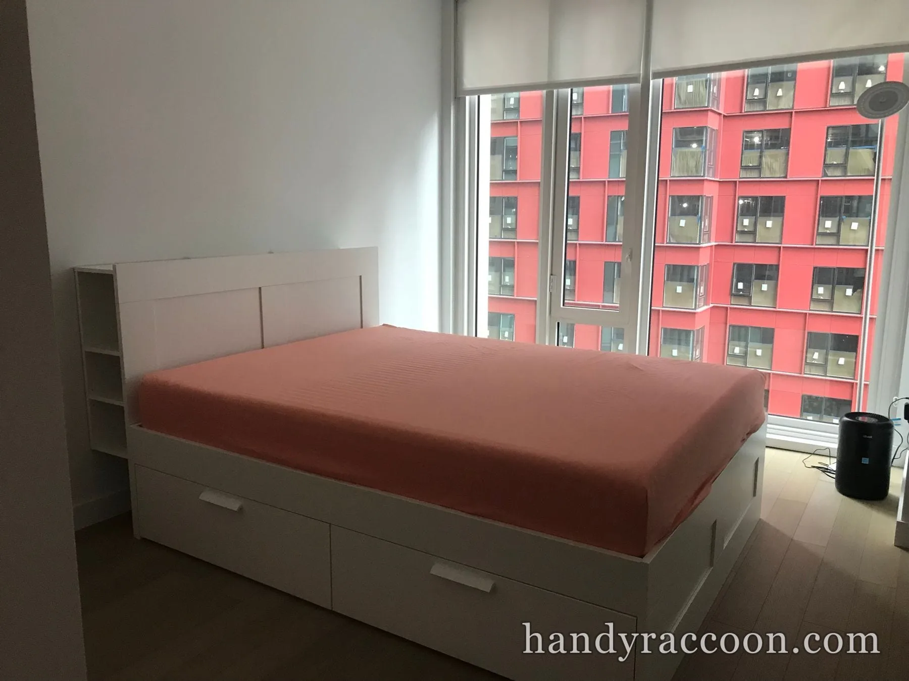 White platform bed with built-in drawers and fitted sheet beside a floor-to-ceiling bedroom window.