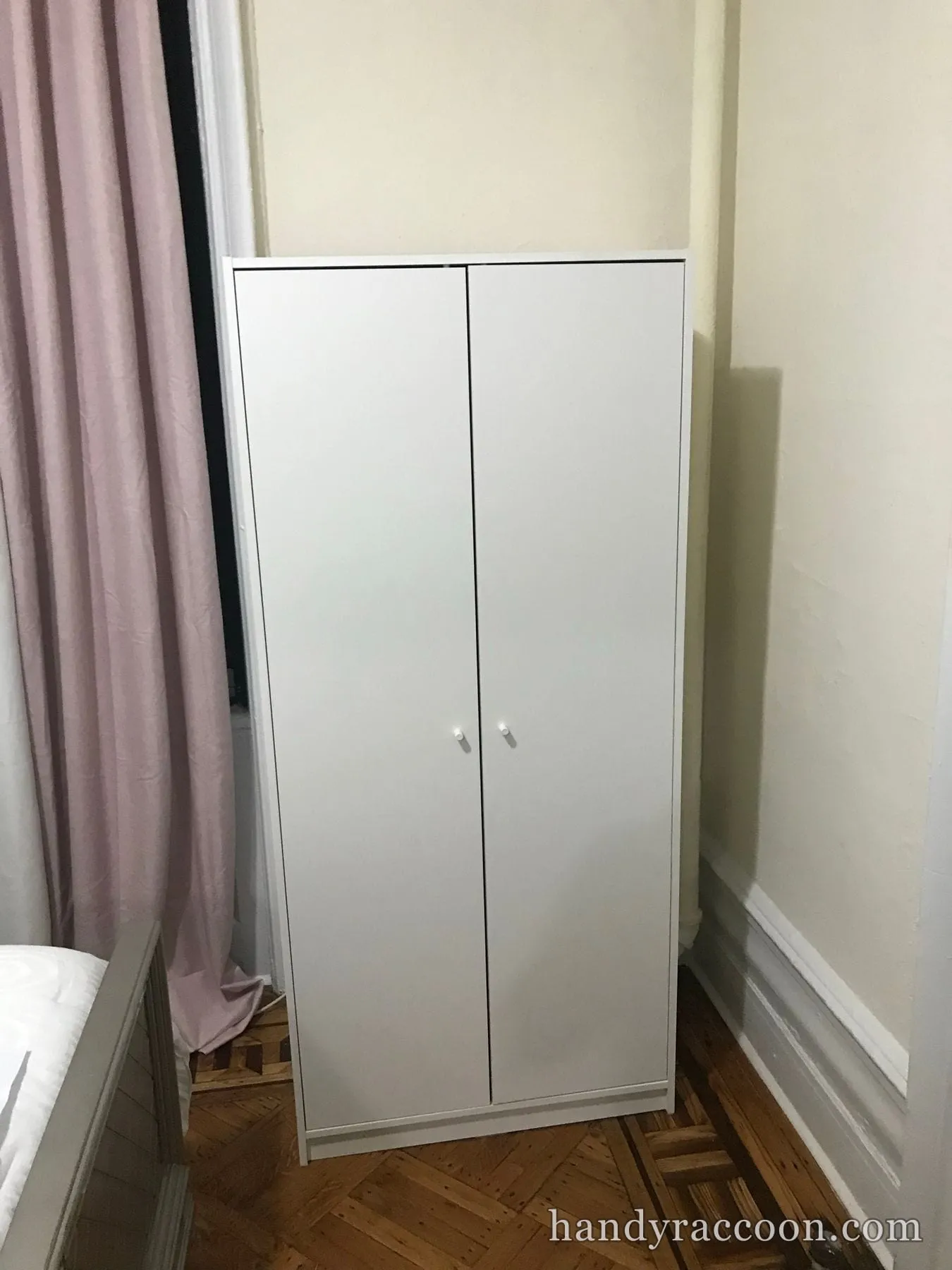 White two-door wardrobe assembled in a bedroom corner beside a pink curtain.