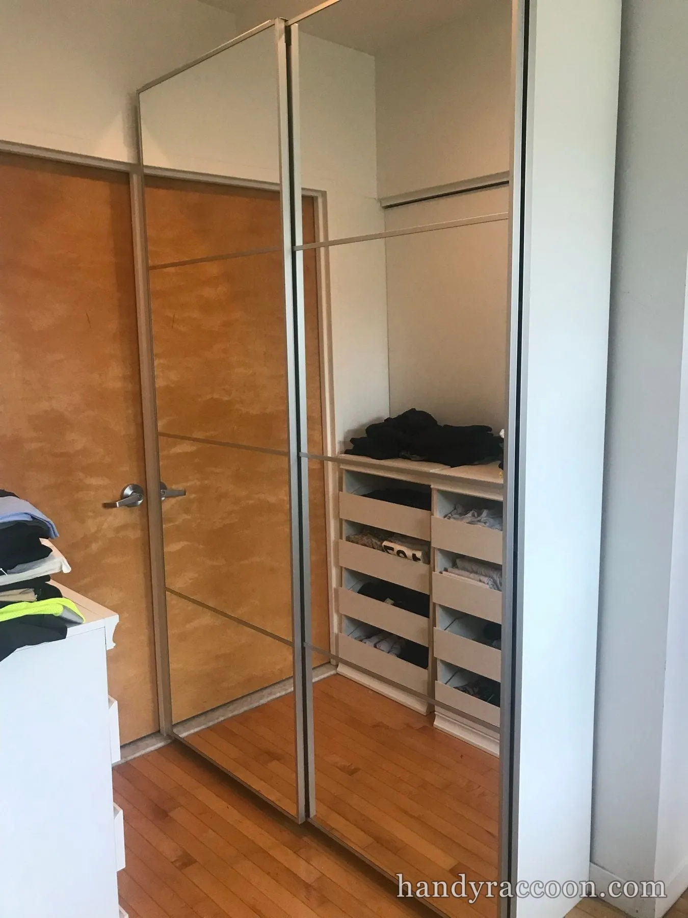 Tall wardrobe with mirrored sliding doors open to reveal pull-out drawers of folded clothes in a bedroom.