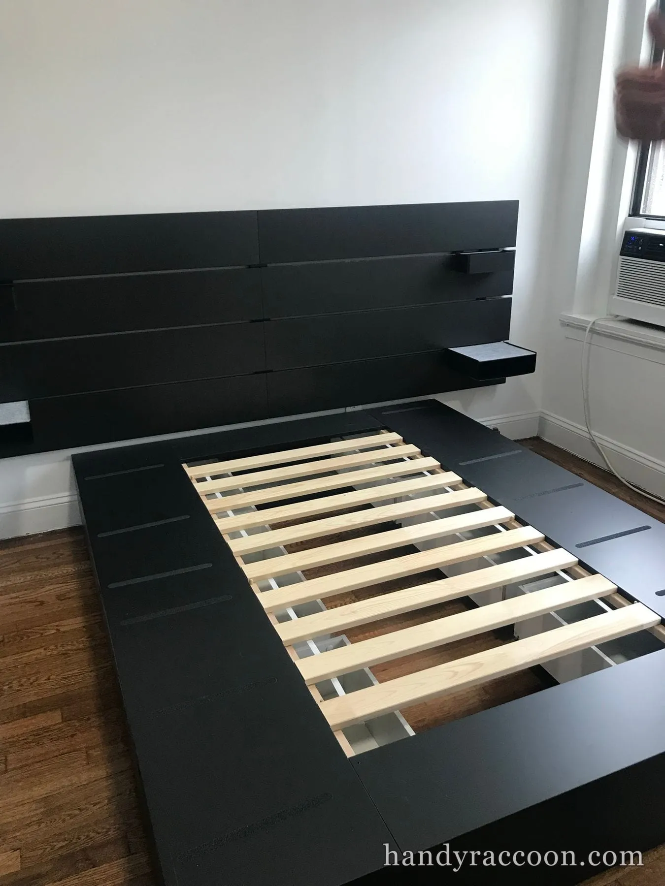 Assembled black platform bed frame with wooden slats and a wall-length floating headboard beside a window.