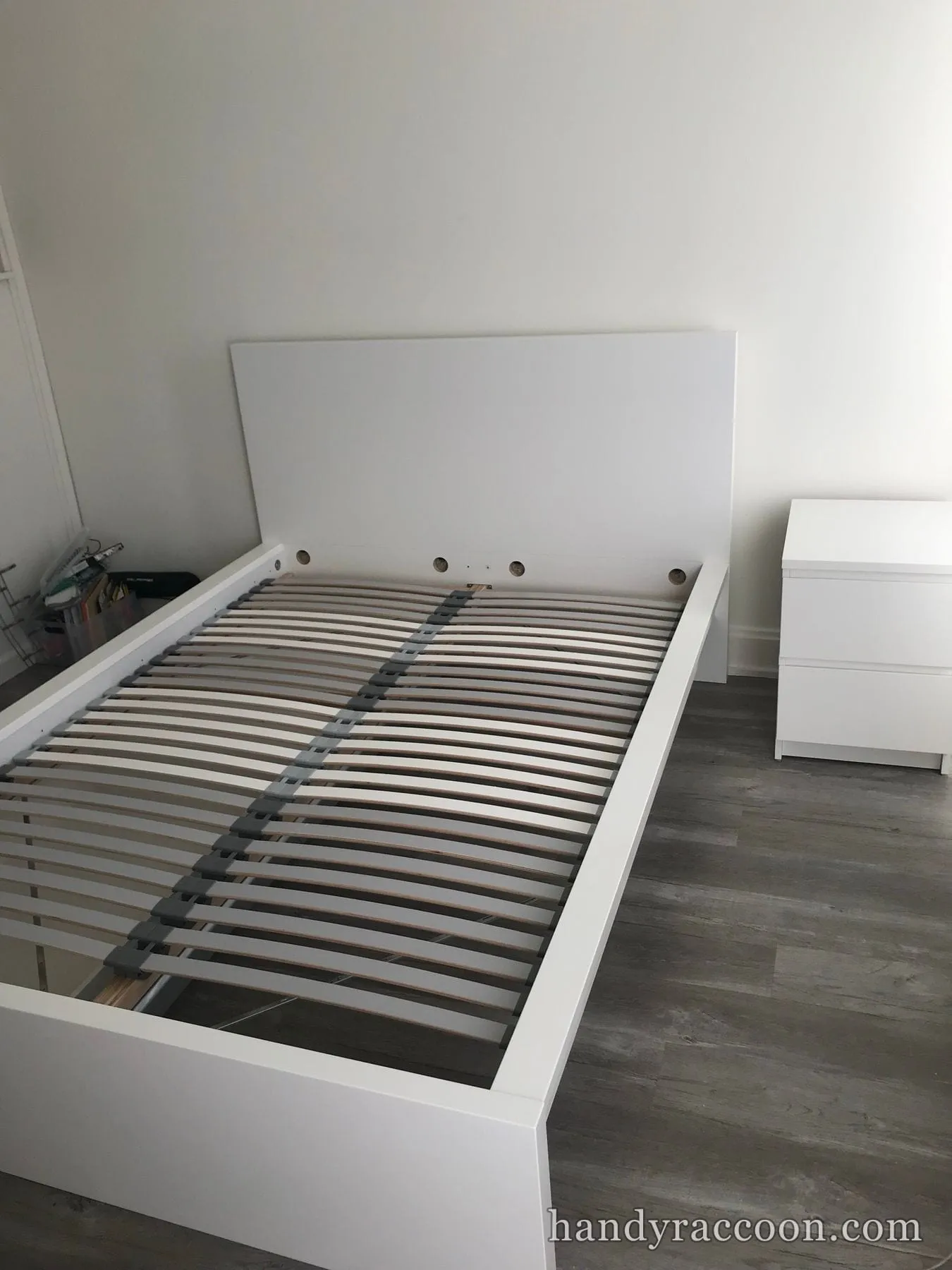 Assembled white bed frame with wooden slats next to a white two-drawer nightstand on a gray floor.