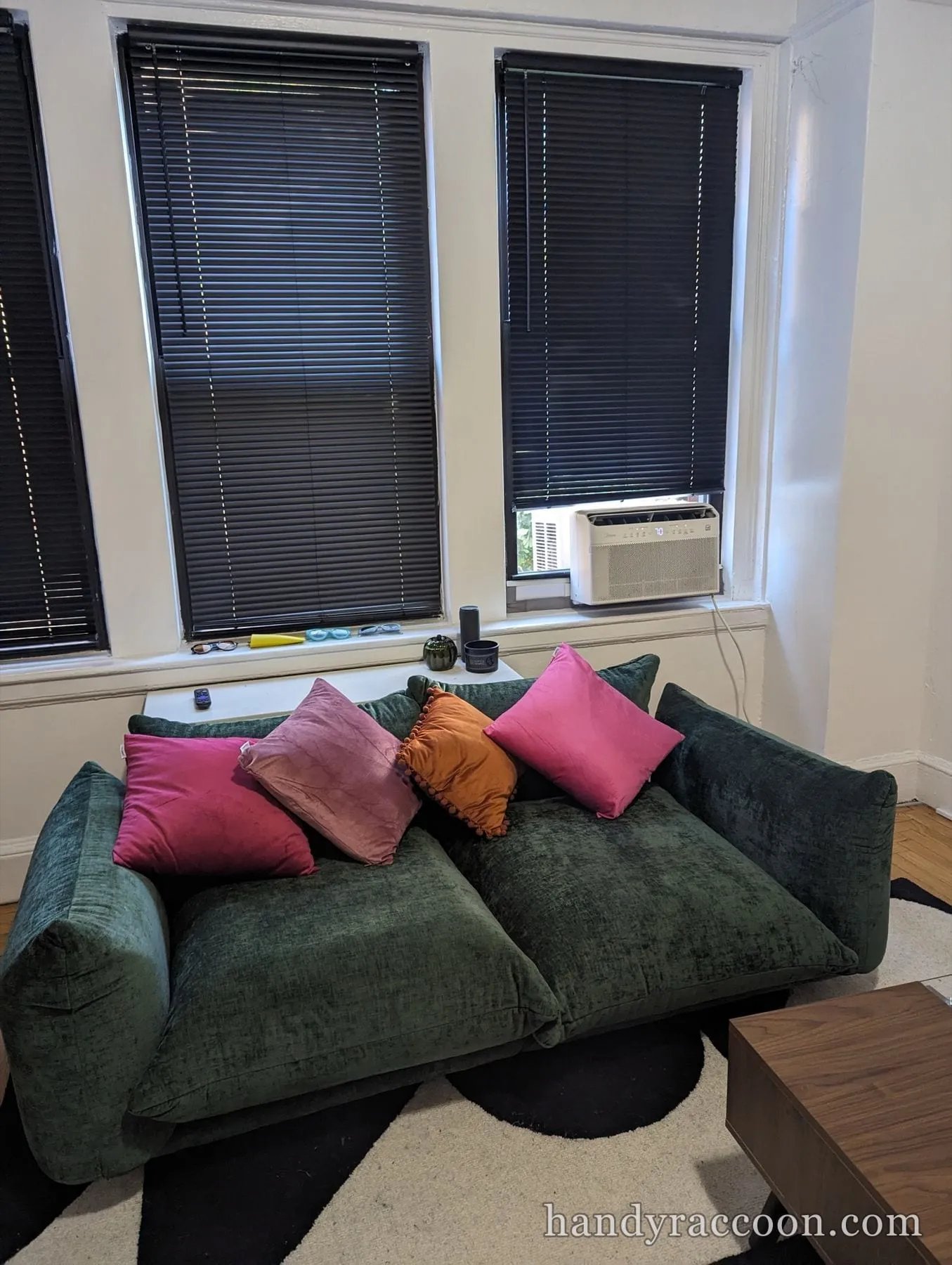 Green velvet loveseat with pink and orange pillows below two windows fitted with dark mini-blinds.