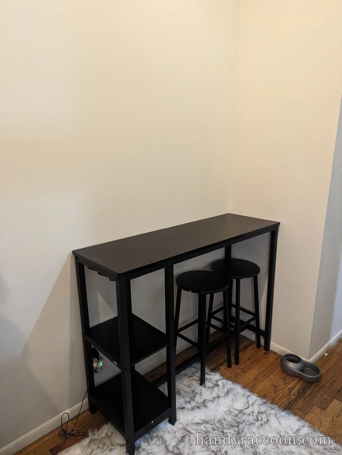 Black narrow bar table with lower shelves and two round stools placed on a rug in a corner.