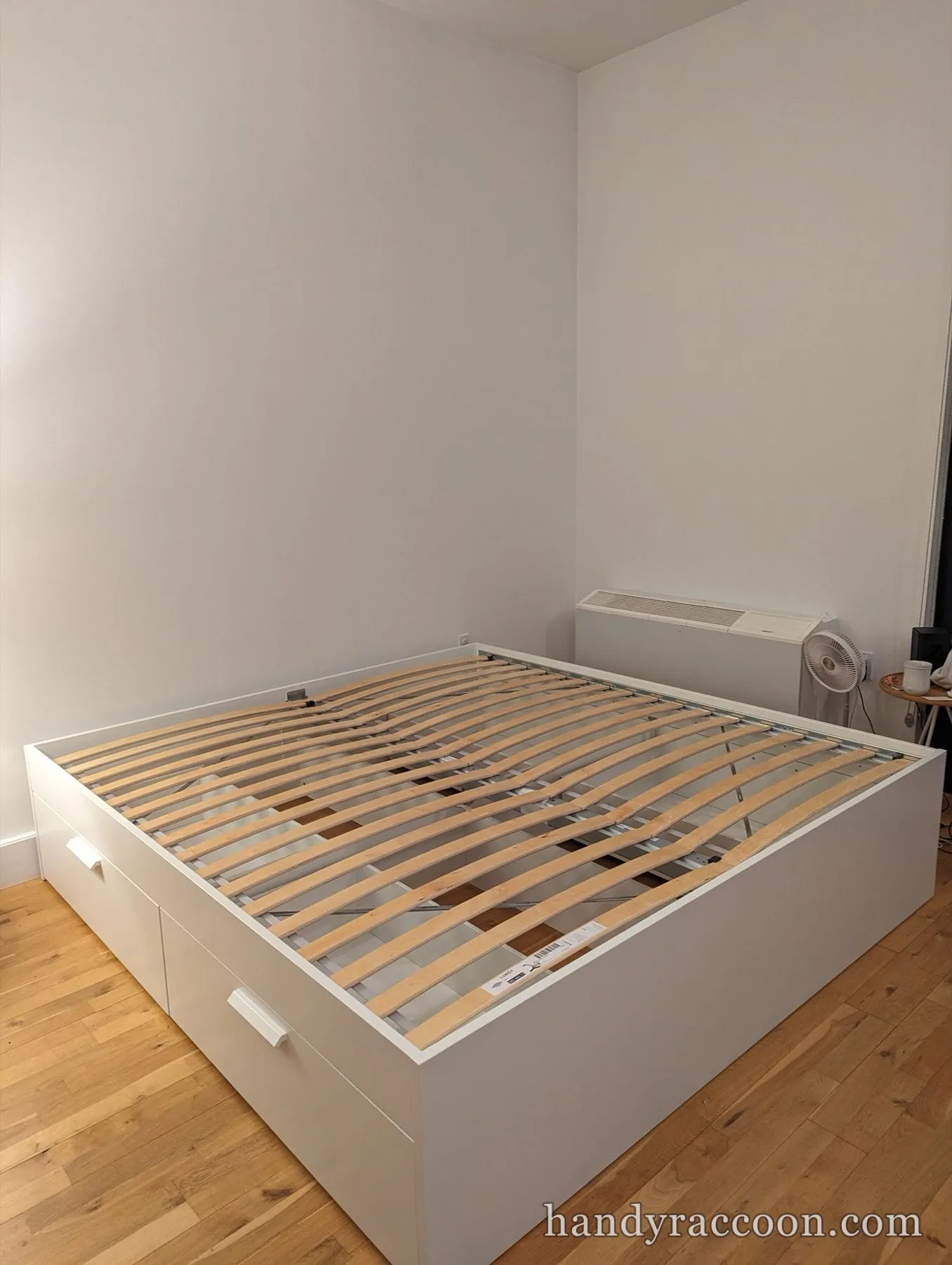 Assembled white platform storage bed with a side drawer and wooden slats on a wood floor.