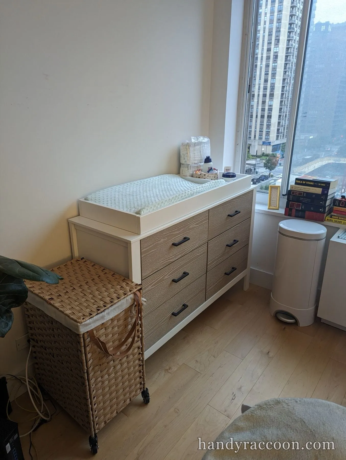 Two-tone nursery dresser with a white changing tray and pad on top, a wicker hamper beside it by a window.