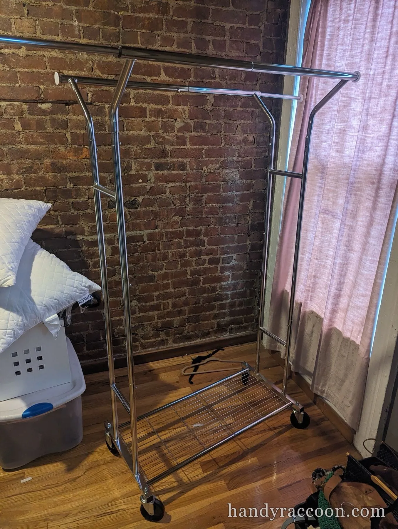 Assembled chrome double-rail rolling garment rack with a lower wire shelf beside an exposed brick wall.