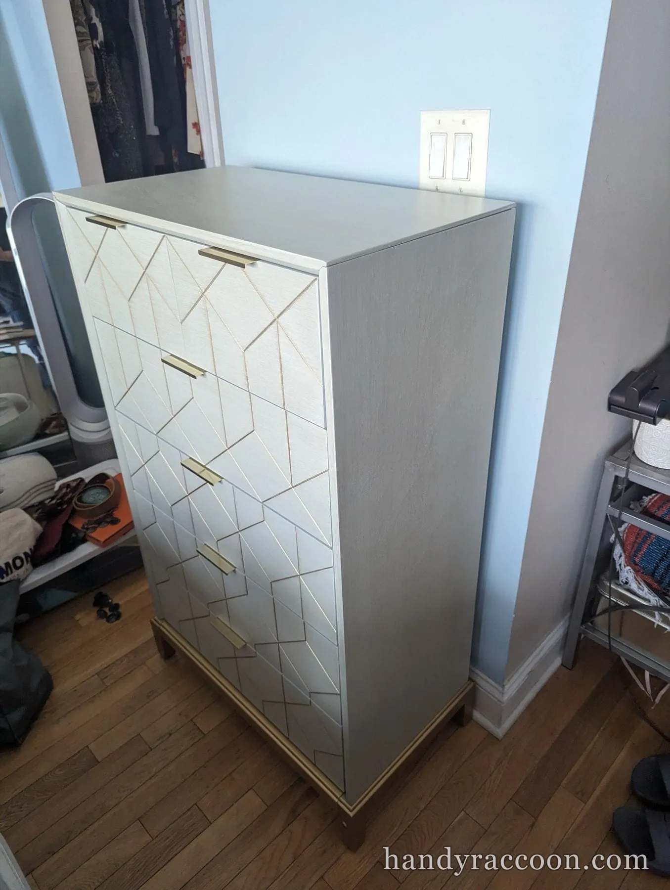 Tall gray chest of drawers with gold geometric inlay and brass handles assembled against a pale blue wall.