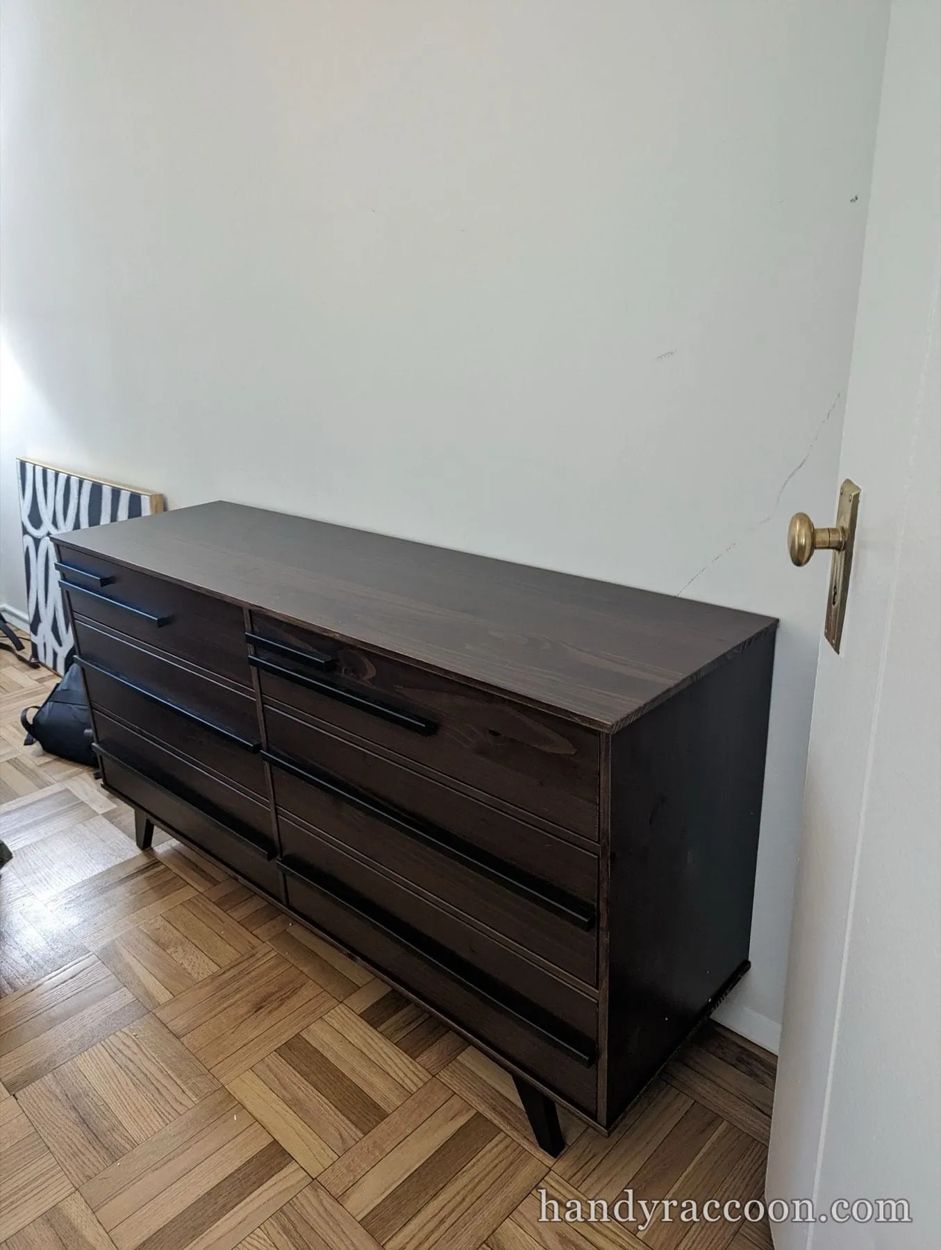 Dark brown wood six-drawer dresser with black bar handles on parquet floor by a white wall.