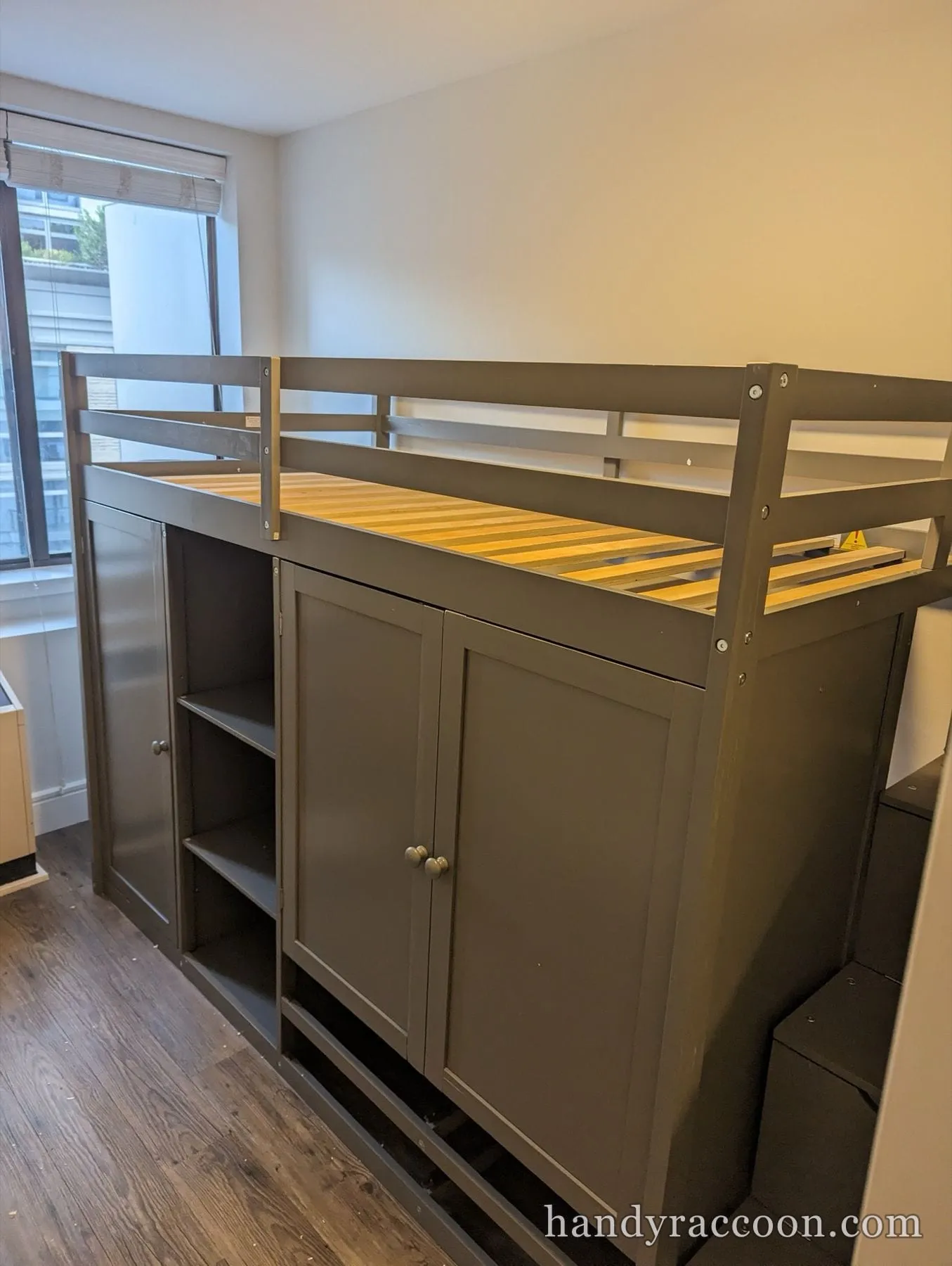 Gray wooden loft bed with storage cabinets and a slatted top beside a bright window.