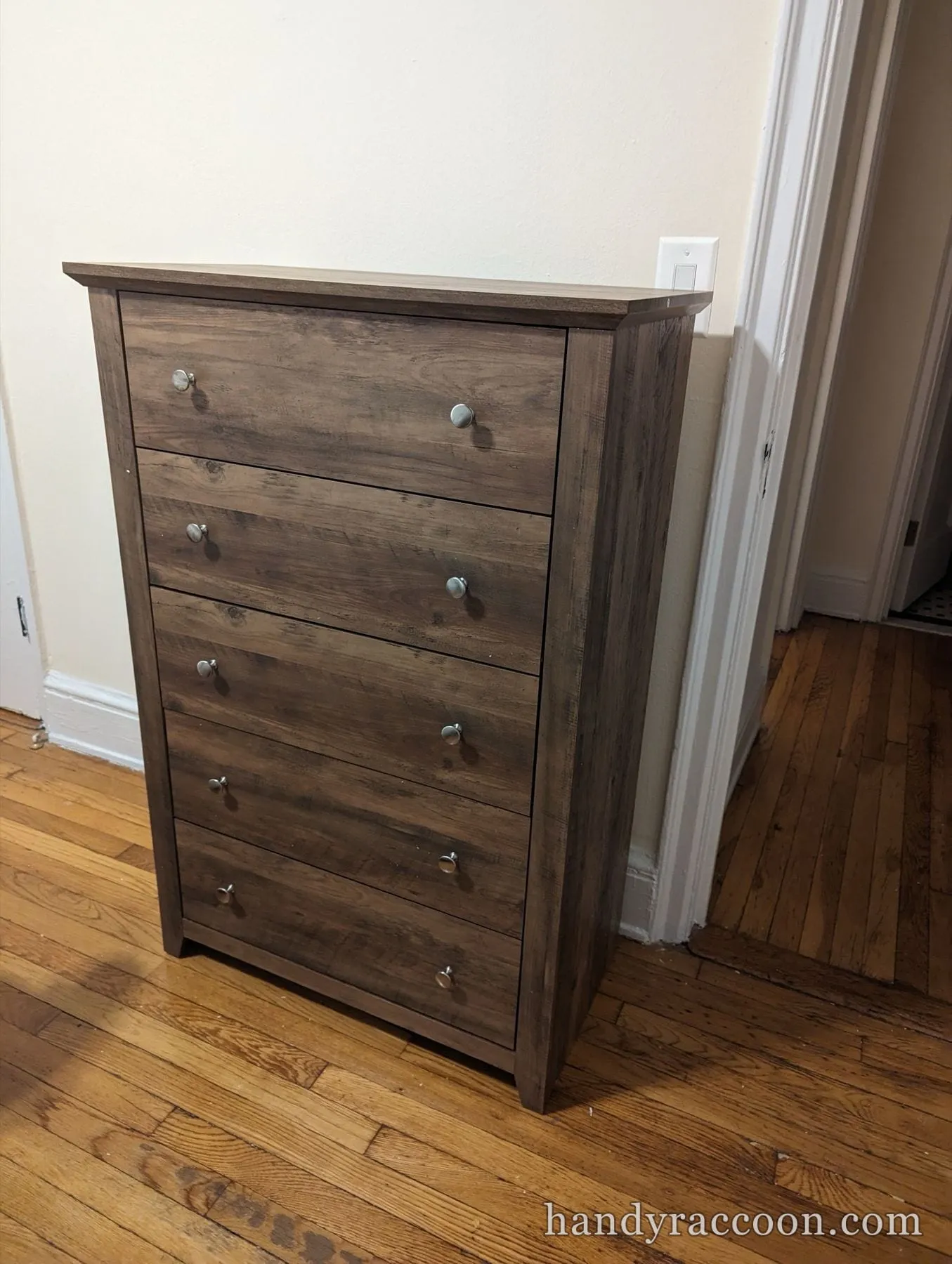 Brown wood-grain five-drawer tall chest with round metal knobs on a hardwood floor.