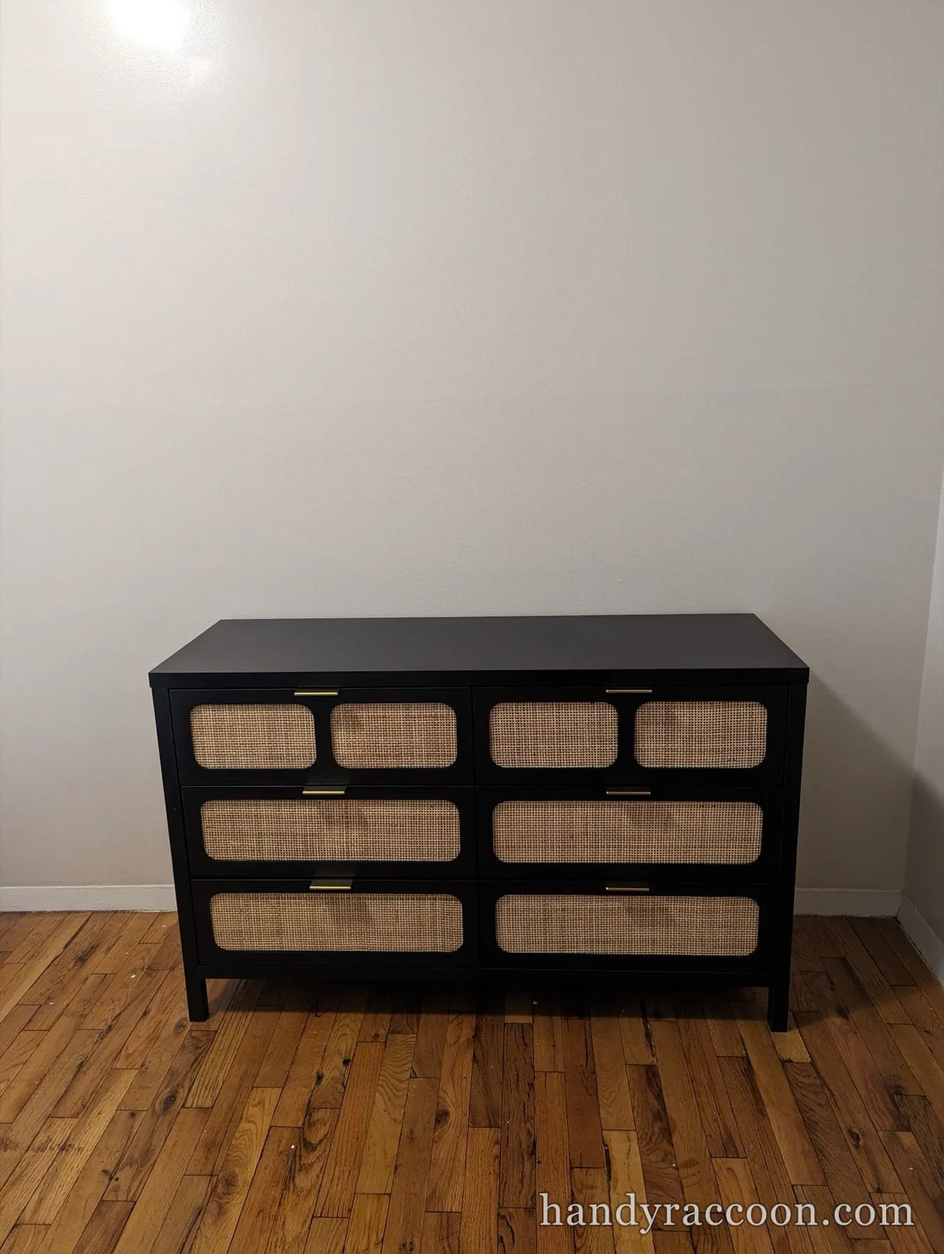 Black dresser with woven cane drawer fronts and gold handles against a gray wall.