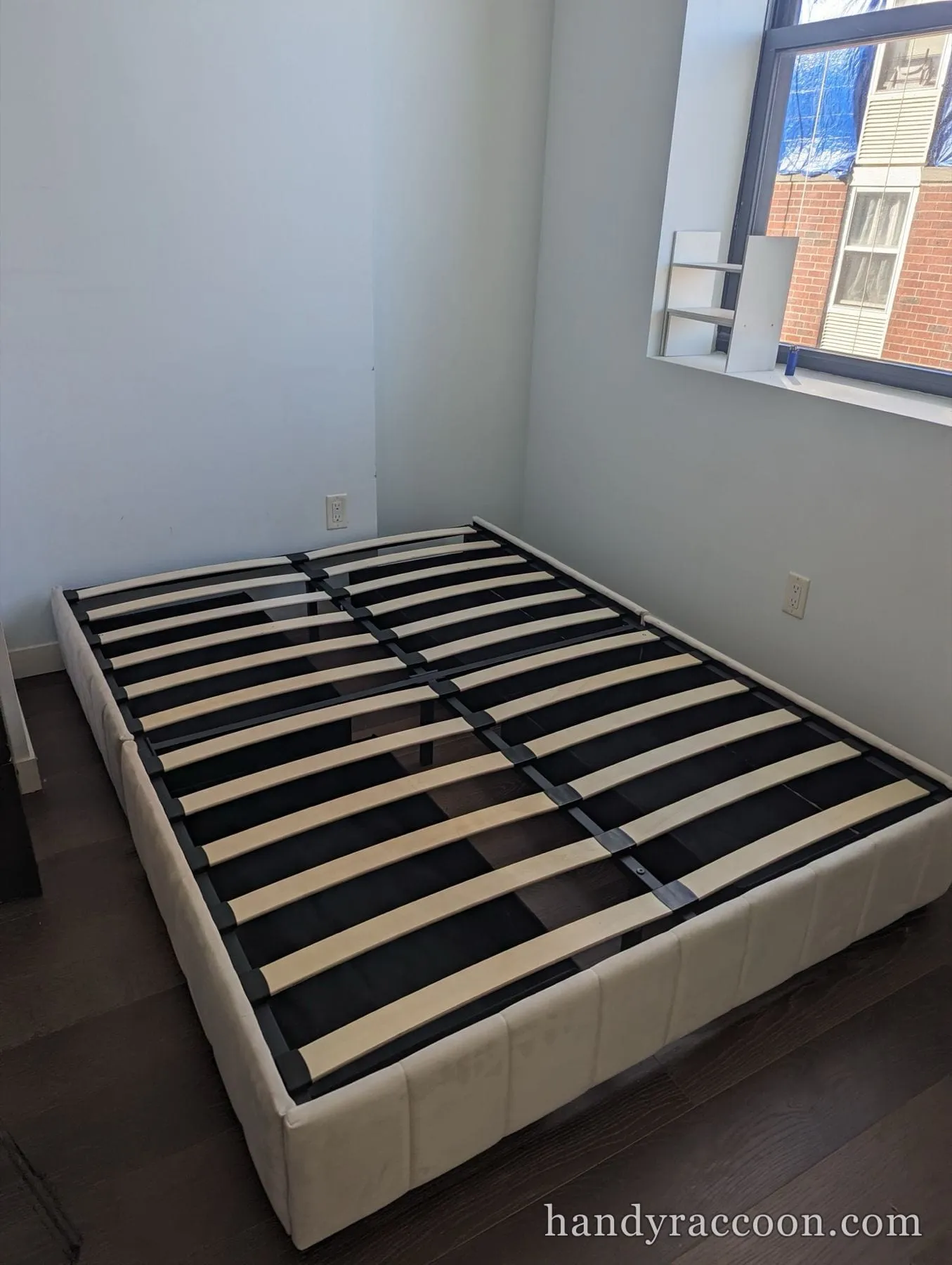 Cream upholstered platform bed base with wooden slats in a corner by a window.
