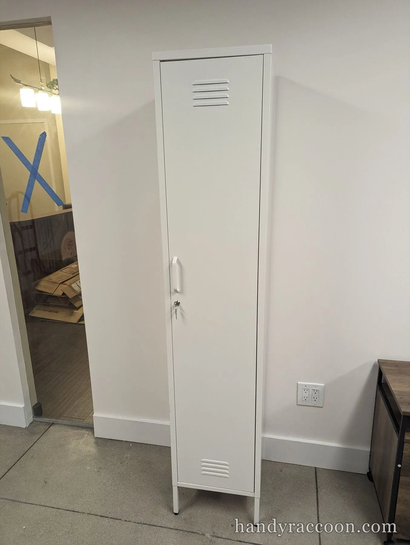 Tall narrow white metal locker with a vented, locking door standing in a corner.