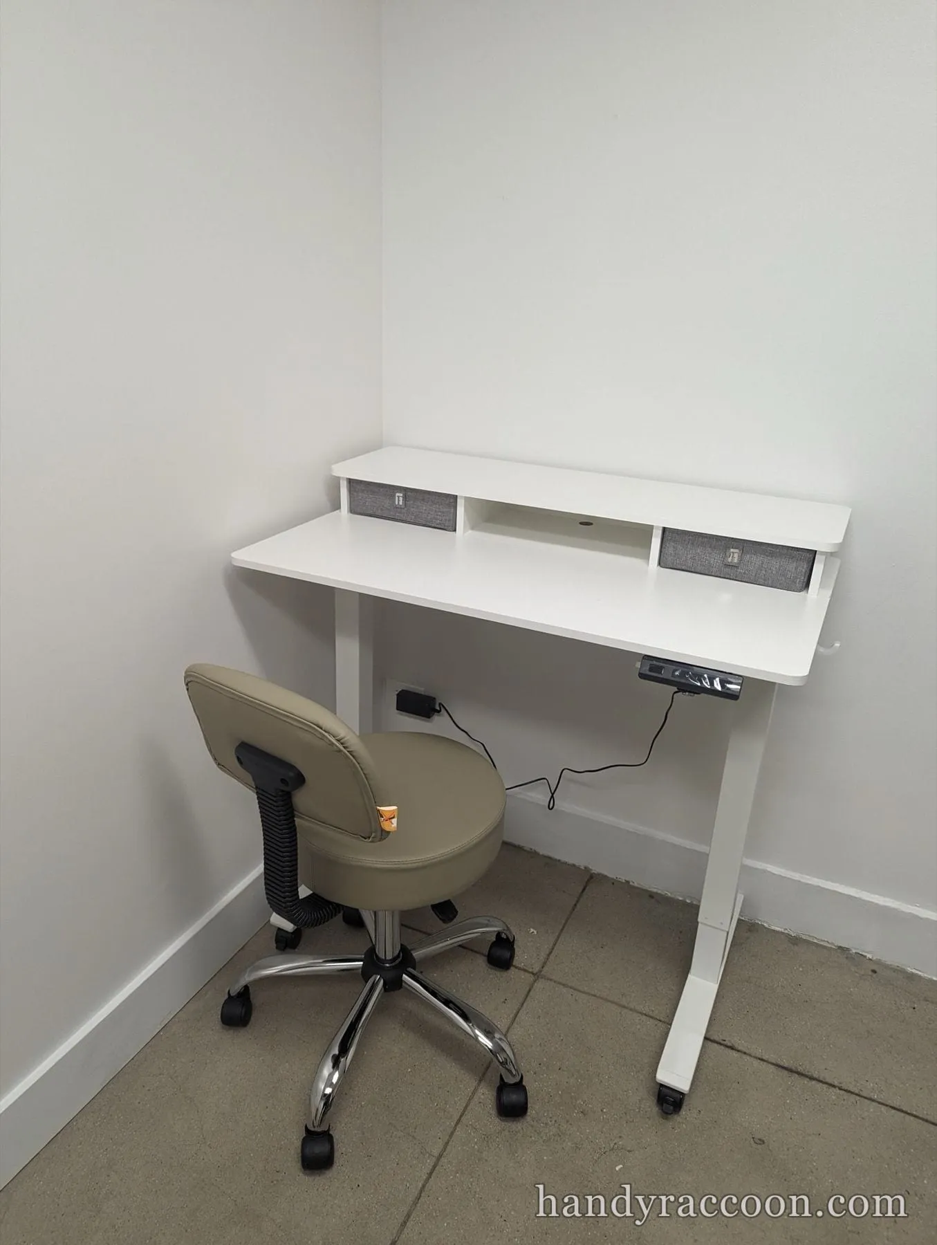 White standing desk with a two-drawer riser and a tan rolling stool in a corner.