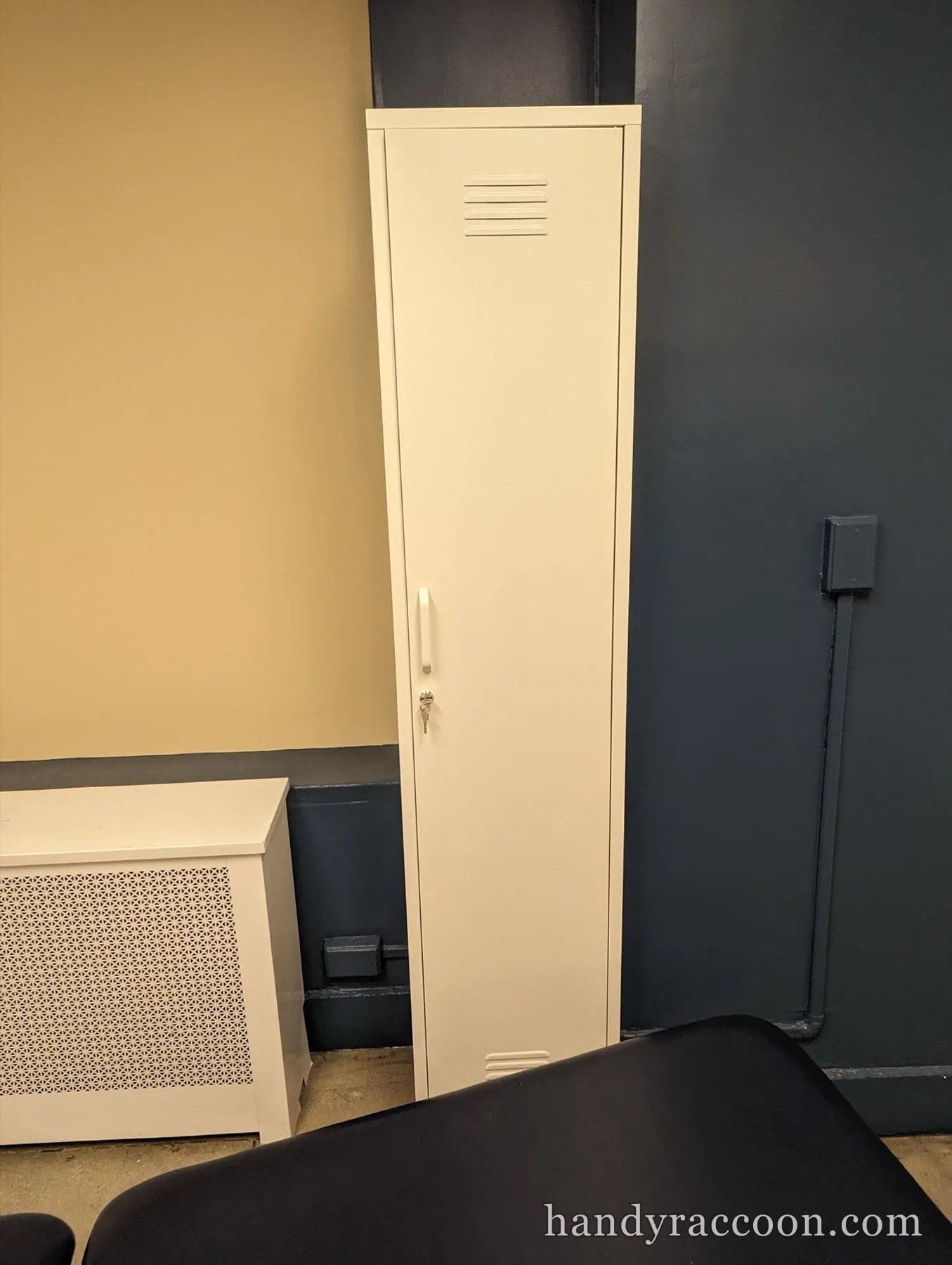 Cream single-door metal locker with a key lock standing in a corner beside a radiator cover.