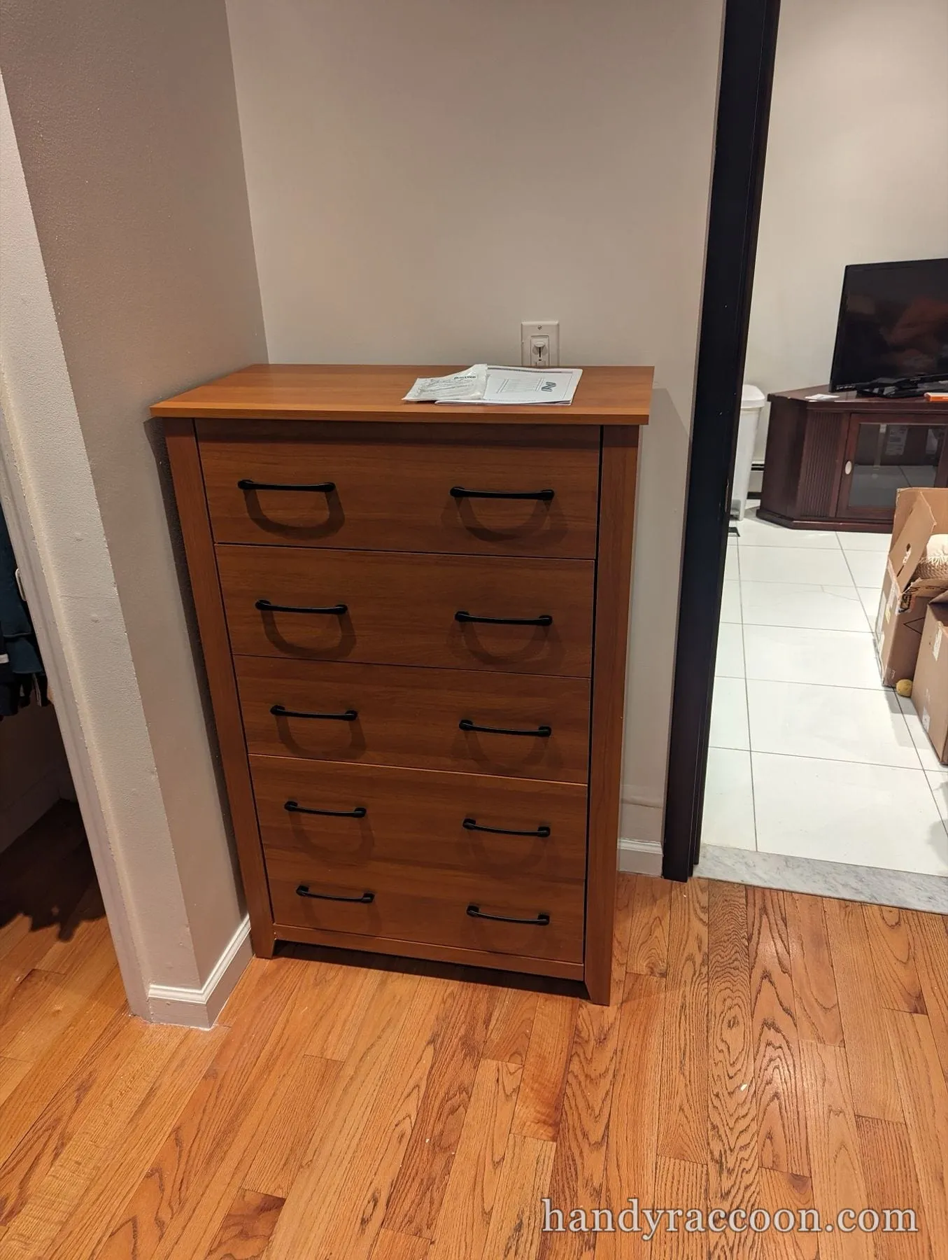 Medium brown five-drawer dresser with black handles against a wall on a hardwood floor.
