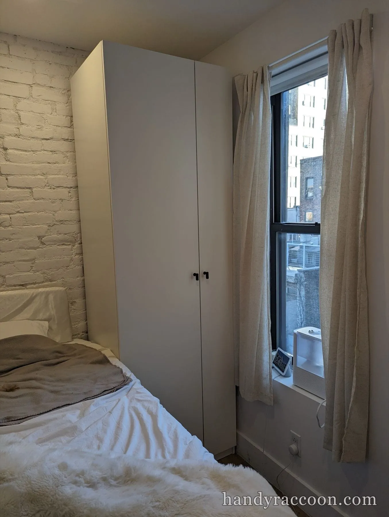 Floor-to-ceiling white two-door wardrobe next to a bed and window in a brick-walled bedroom.