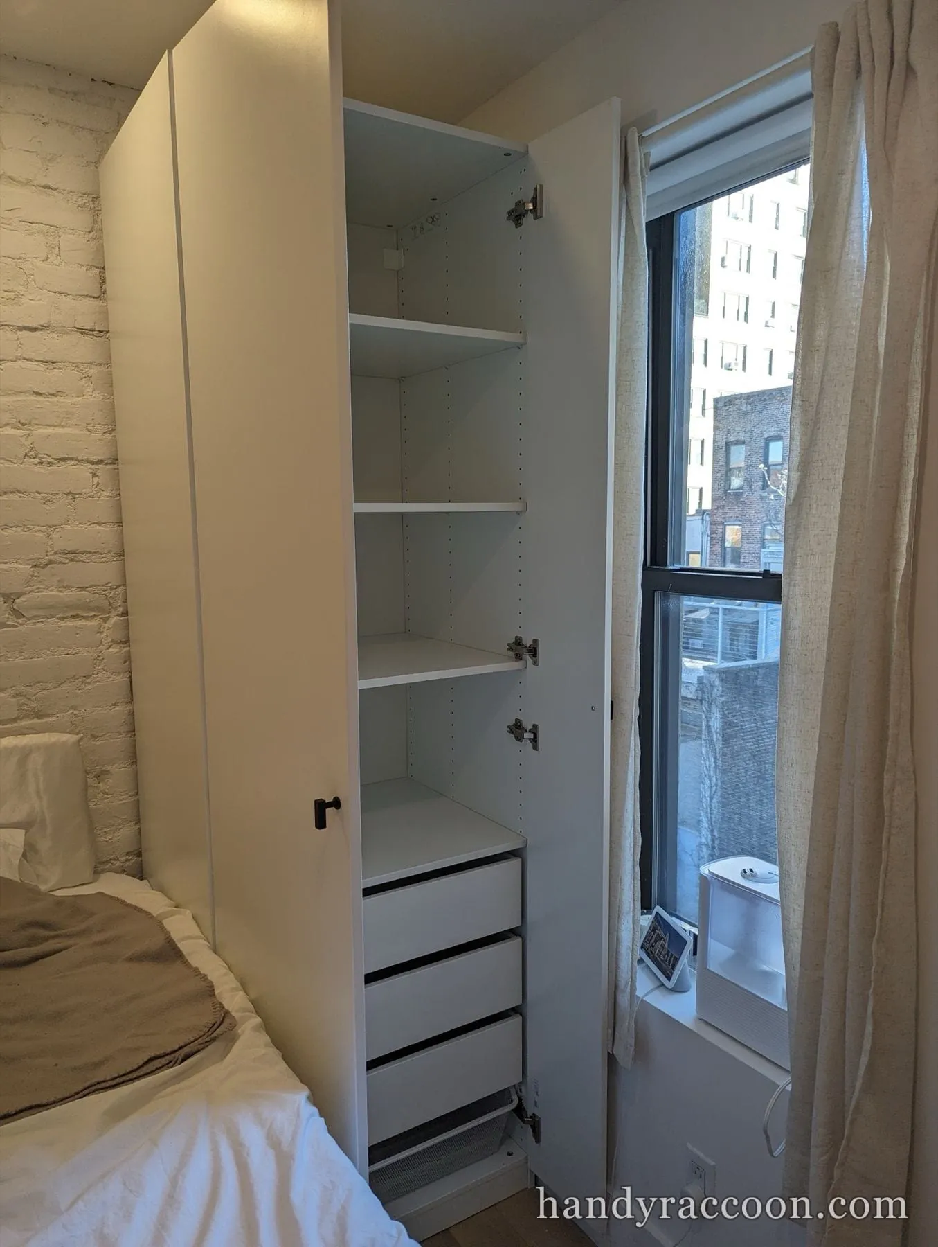 Open white wardrobe showing internal shelves and three stacked drawers in a bedroom.