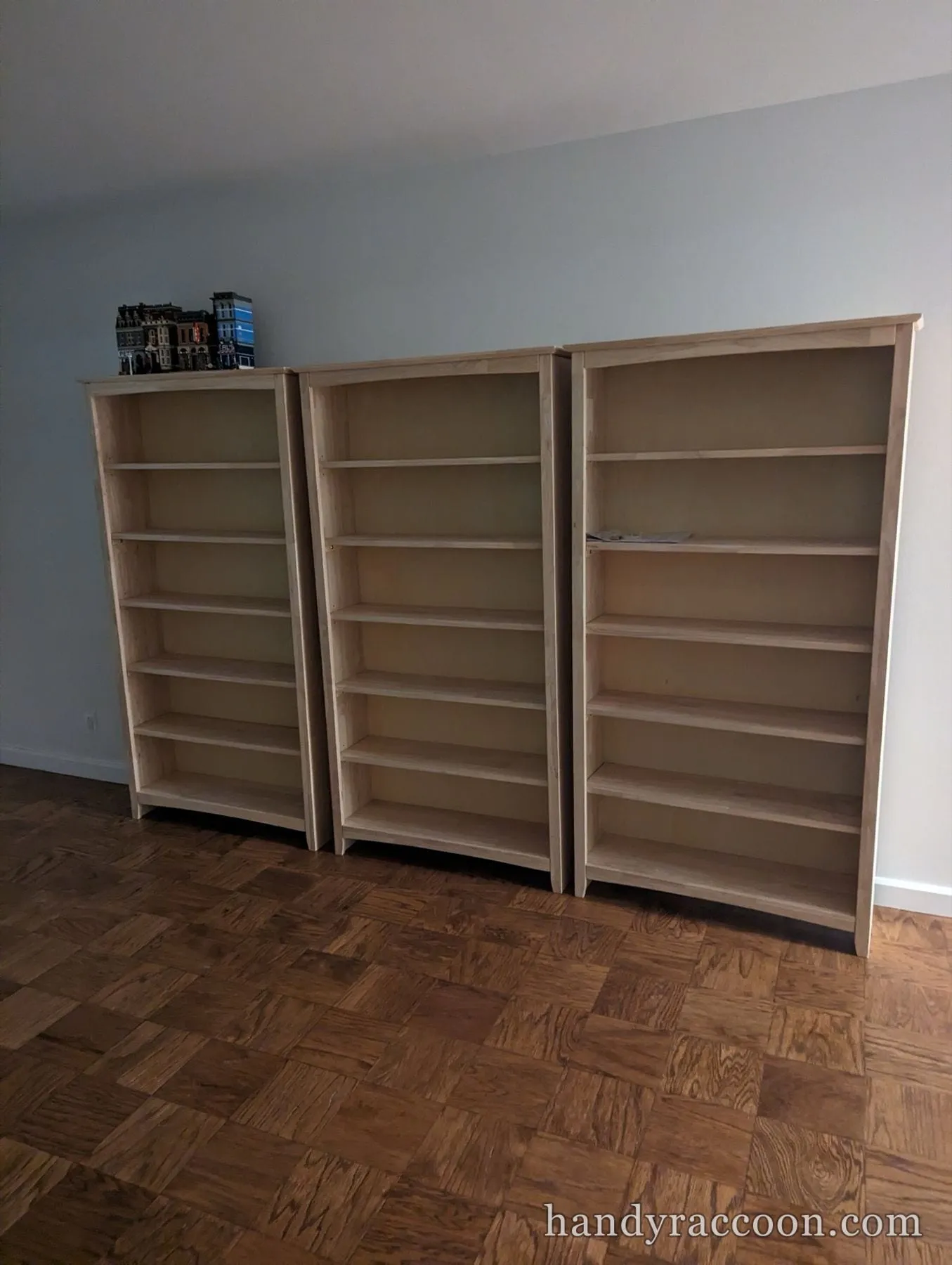 Row of three tall unfinished wood bookcases with empty shelves against a wall.