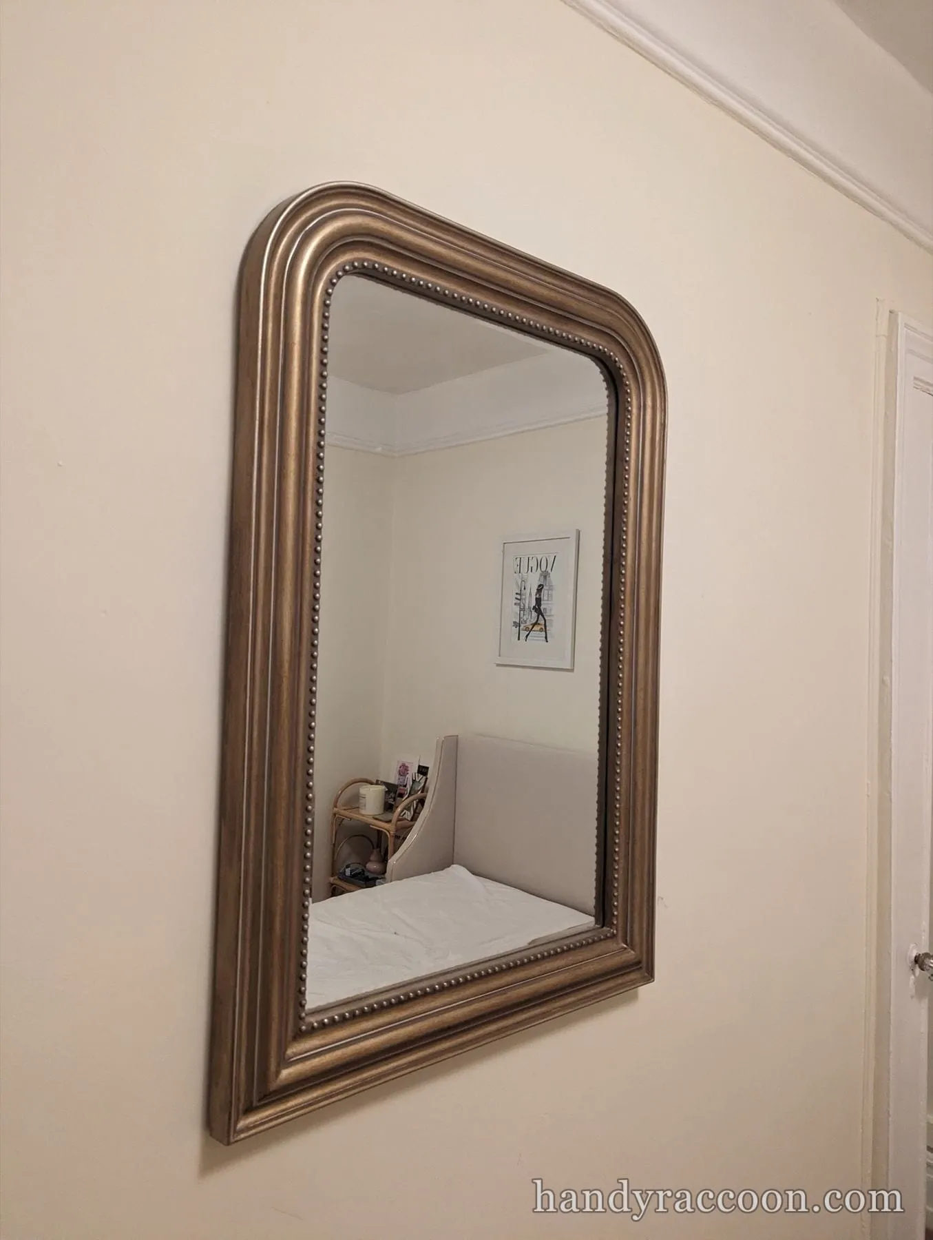 Arch-top mirror with a beaded bronze frame hung on a wall, reflecting a bedroom.