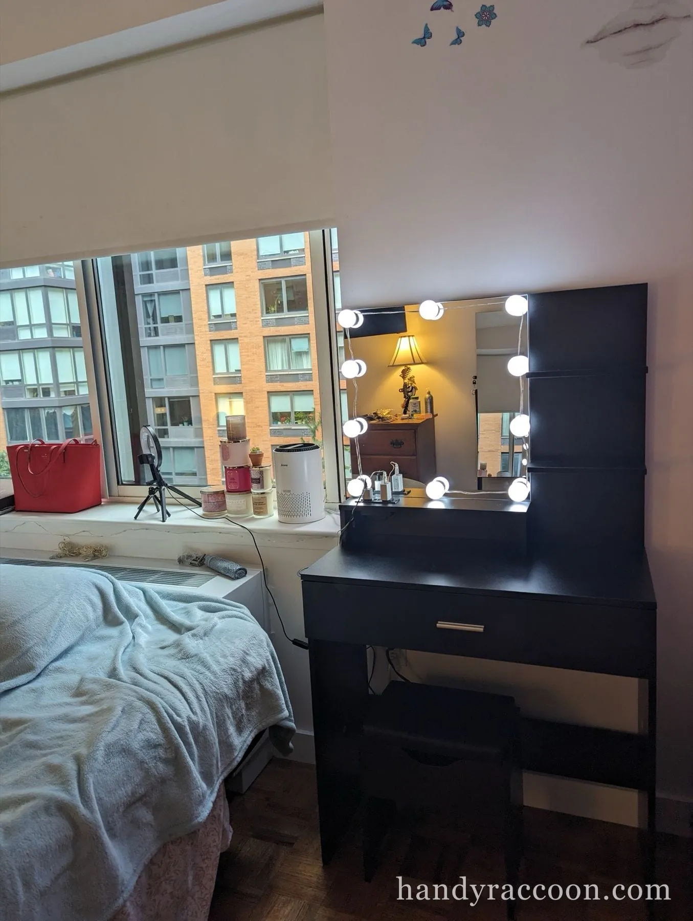 Black vanity desk with a bulb-lit mirror and matching stool set beside a bedroom window.