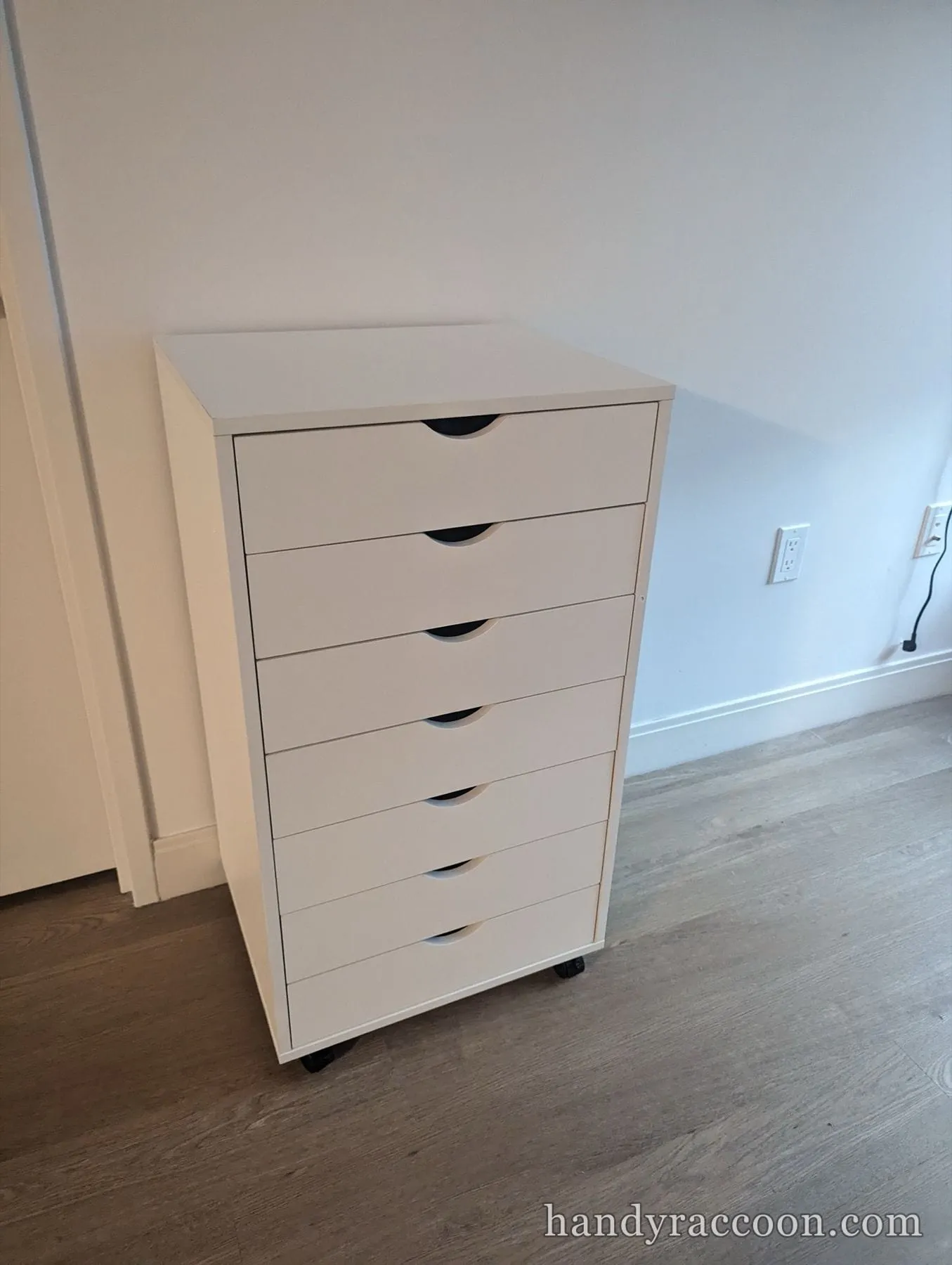 Tall white chest of seven shallow drawers on wheels against a wall.