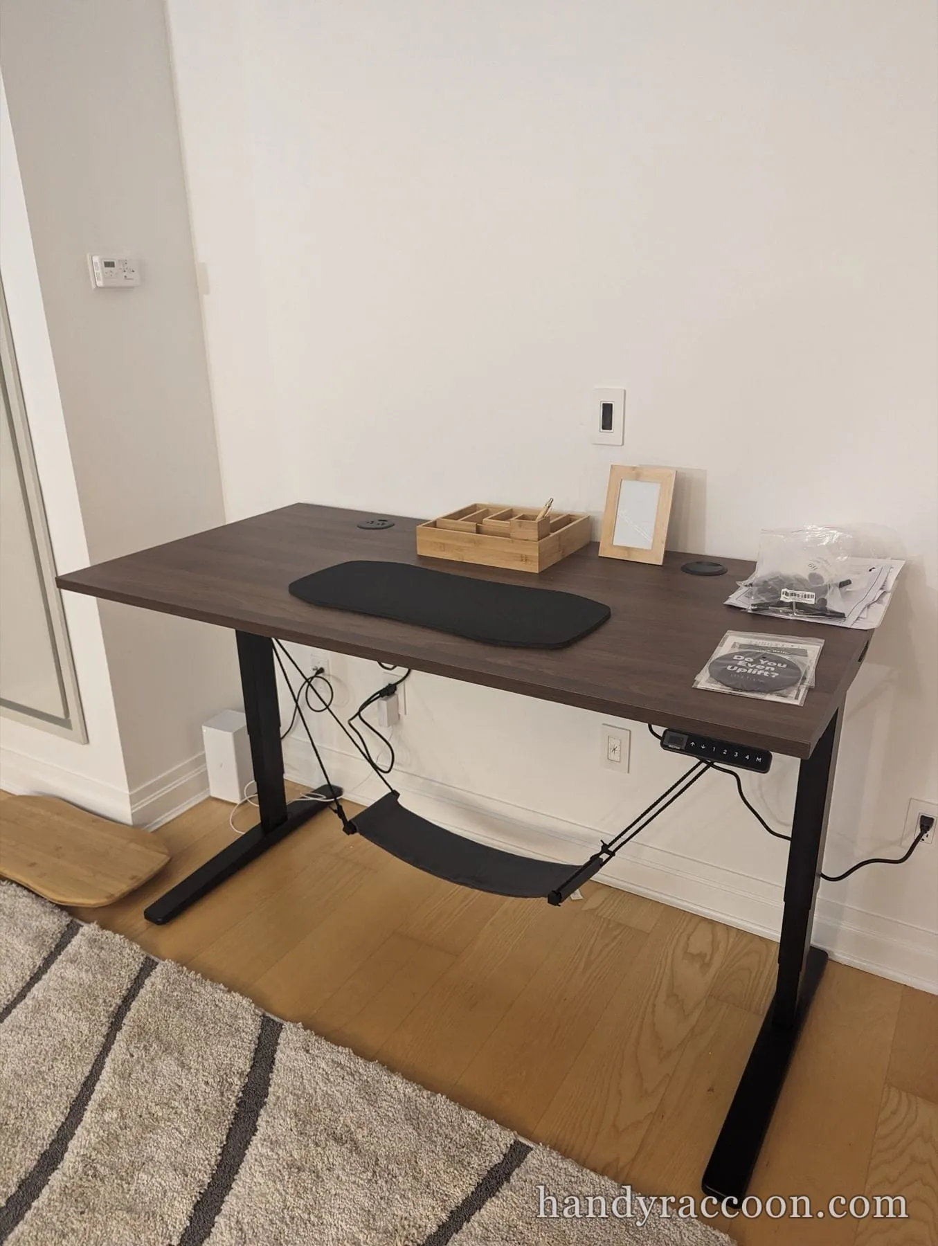 Electric standing desk with a dark wood top, black legs, control panel and under-desk hammock.