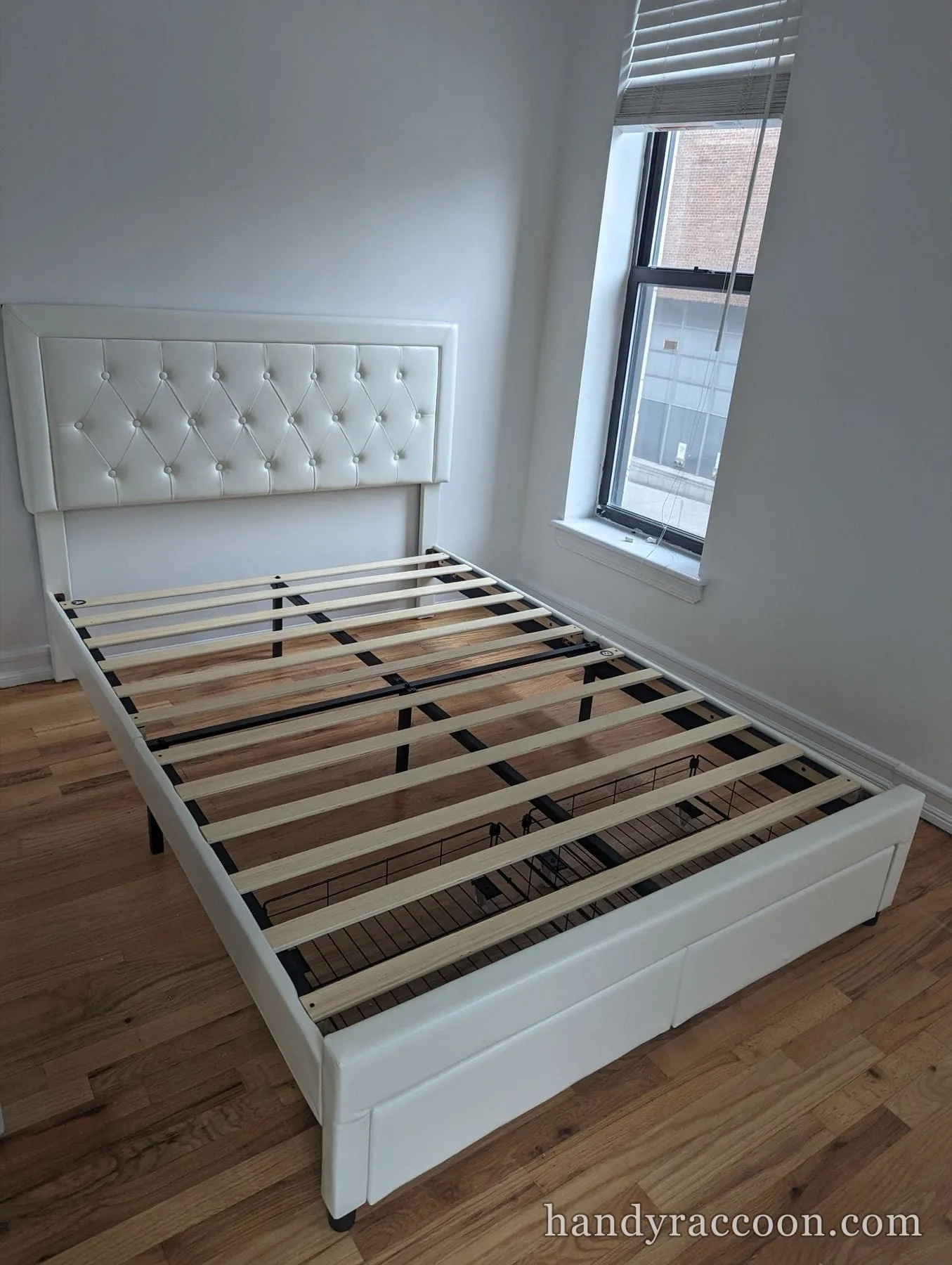 White upholstered platform bed frame with tufted headboard and wood slats on a wood floor.