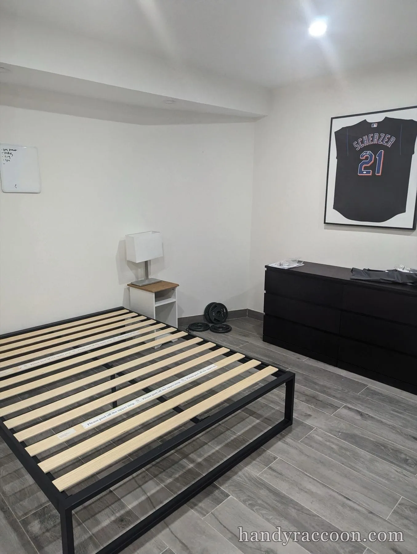 Black metal platform bed frame with light wood slats on gray tile floor near a dresser.