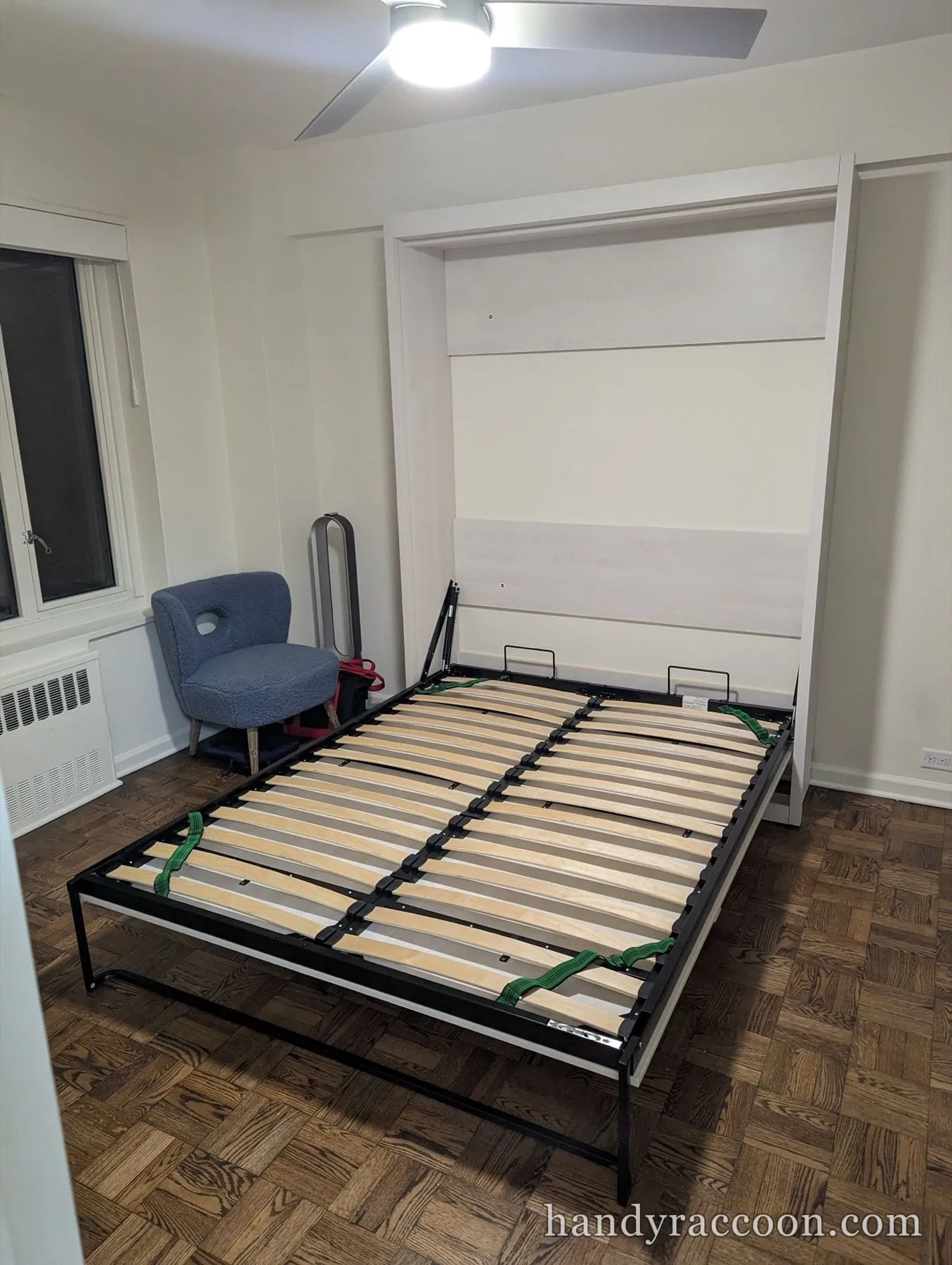 Fold-down Murphy wall bed open with a wood slat frame in a room with parquet floor.