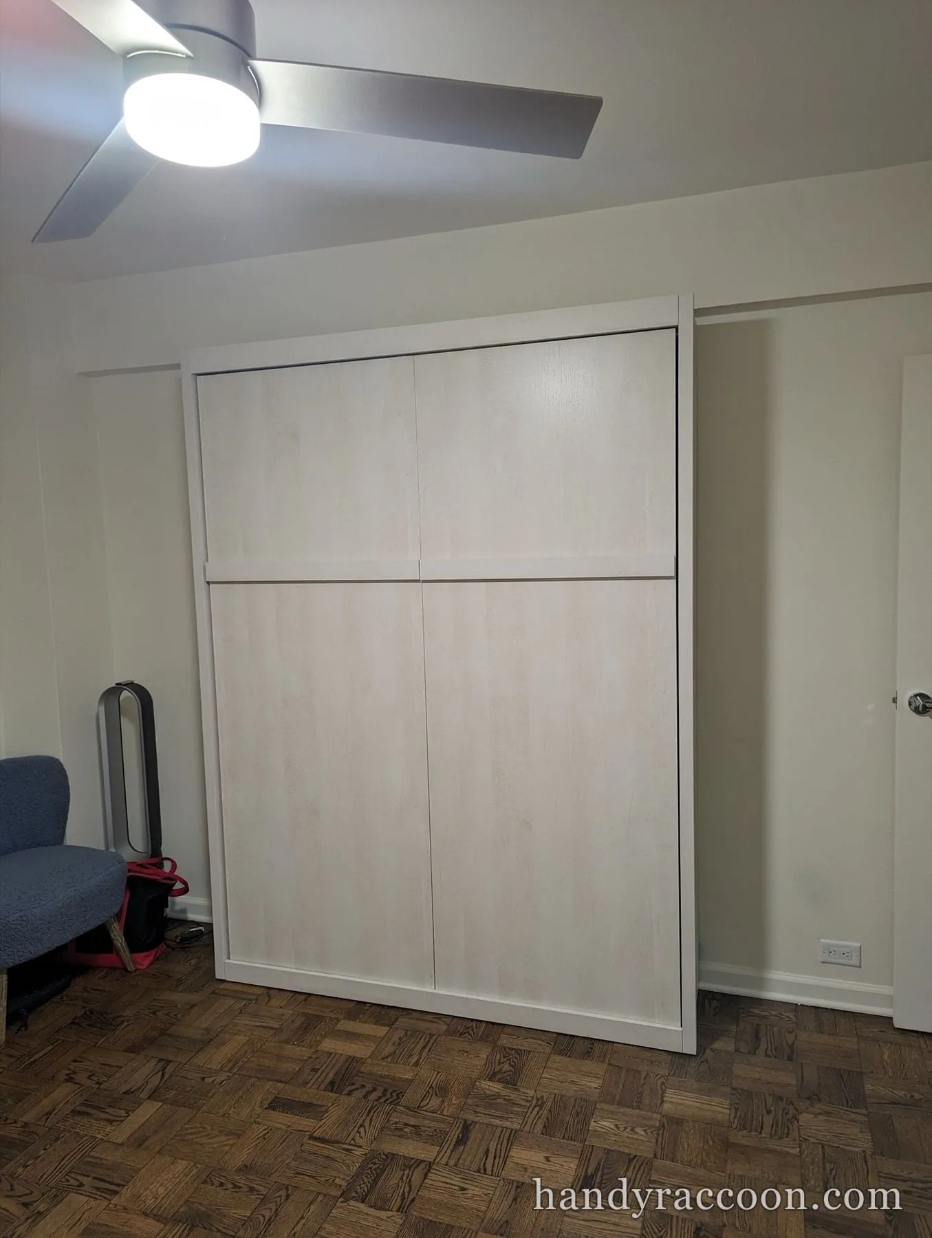 Closed Murphy wall bed cabinet with light panel doors in a room with parquet floor.