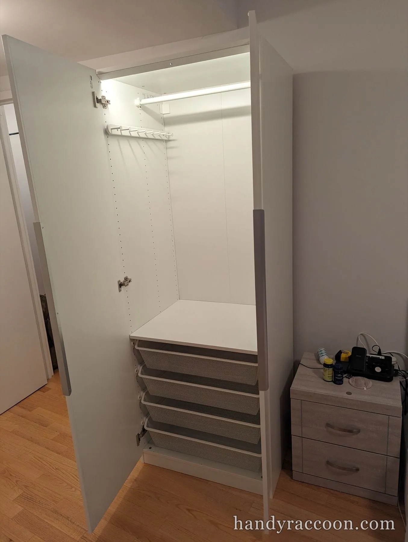 Open white wardrobe lit inside with pull-out mesh baskets and a pants rack, beside a nightstand.
