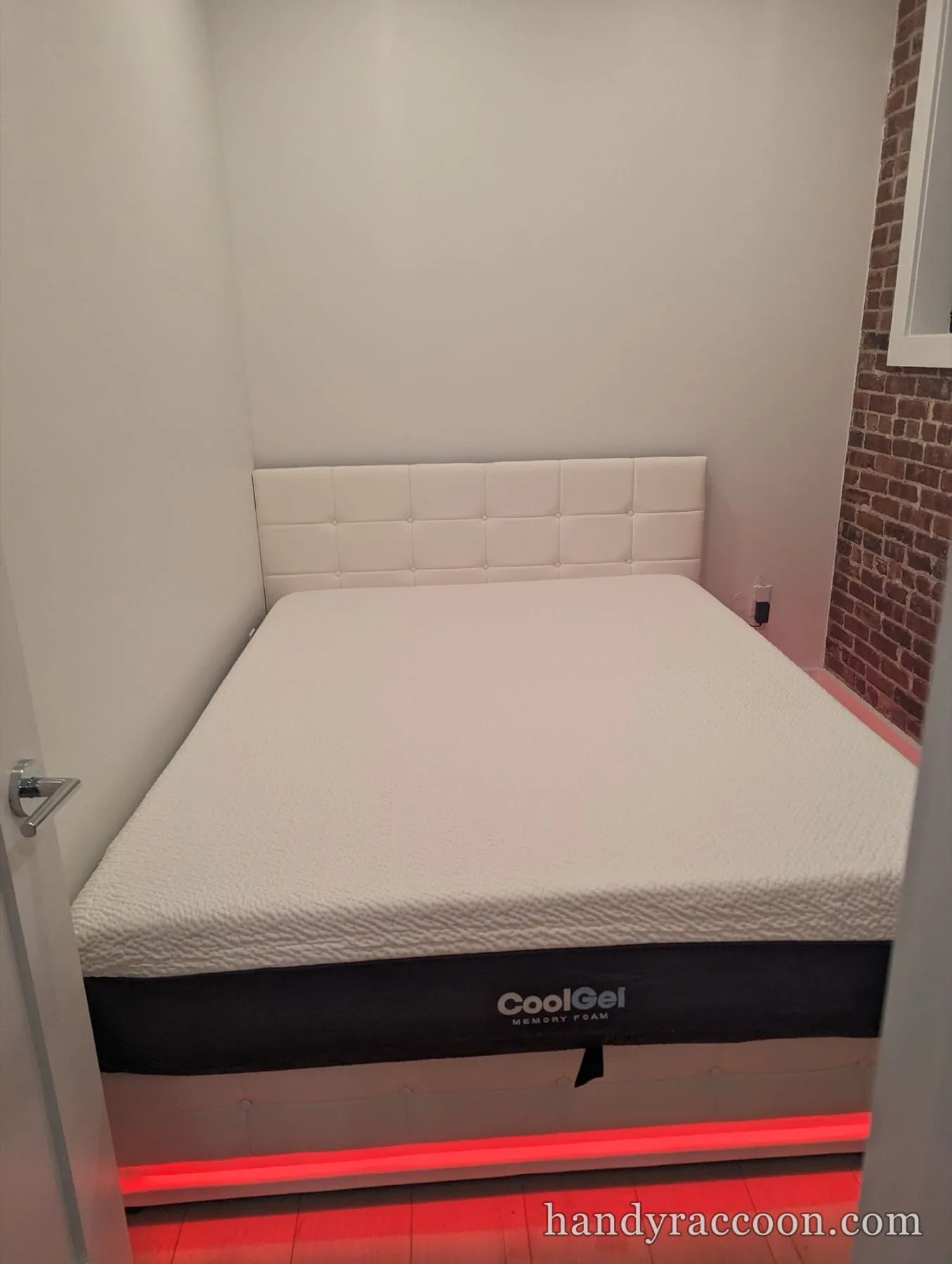 White upholstered bed with a memory foam mattress and red under-bed light near a brick wall.