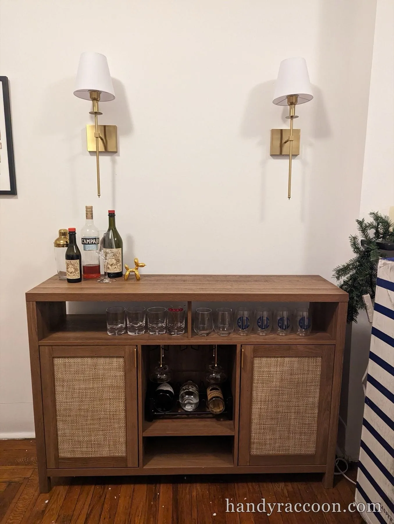 Wood bar cabinet with cane door panels, stemware and bottles below two wall sconces.
