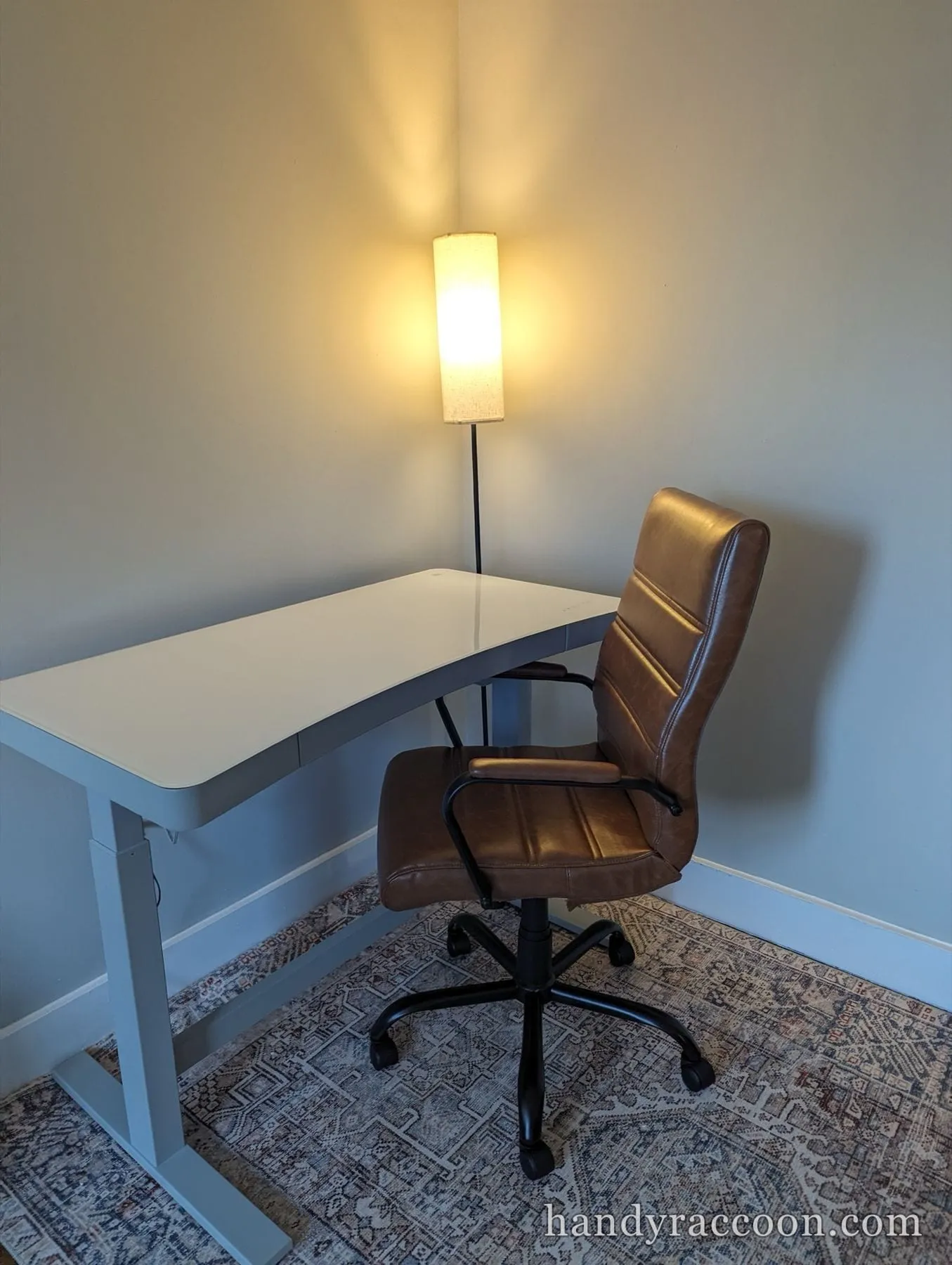 White adjustable standing desk with a brown leather office chair on a patterned rug.