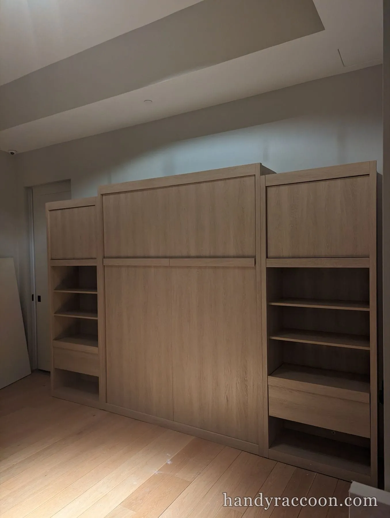Light oak Murphy bed cabinet flanked by open shelving units, closed against the wall.