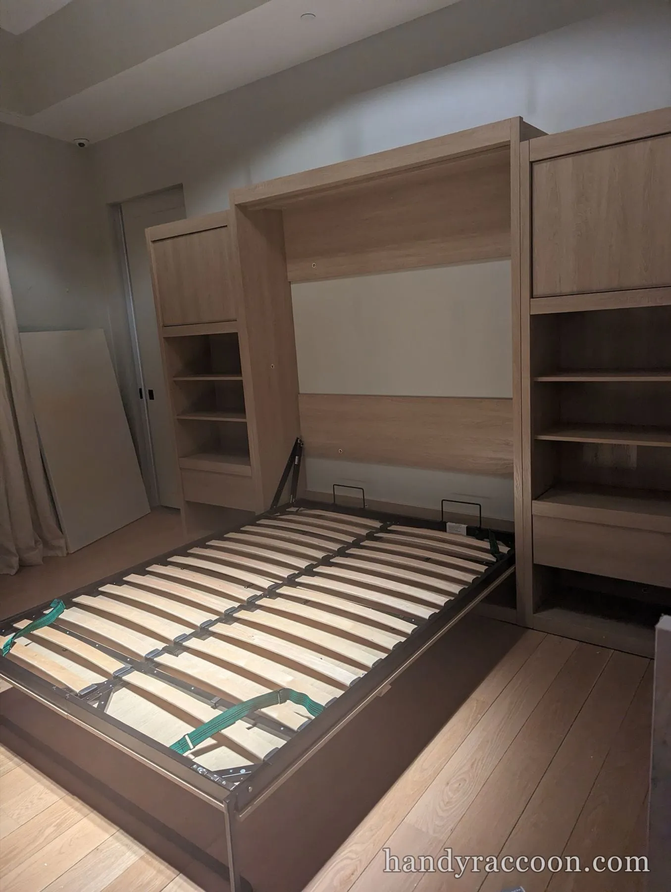 Light oak Murphy bed open with a slat frame between two shelving towers on a wood floor.