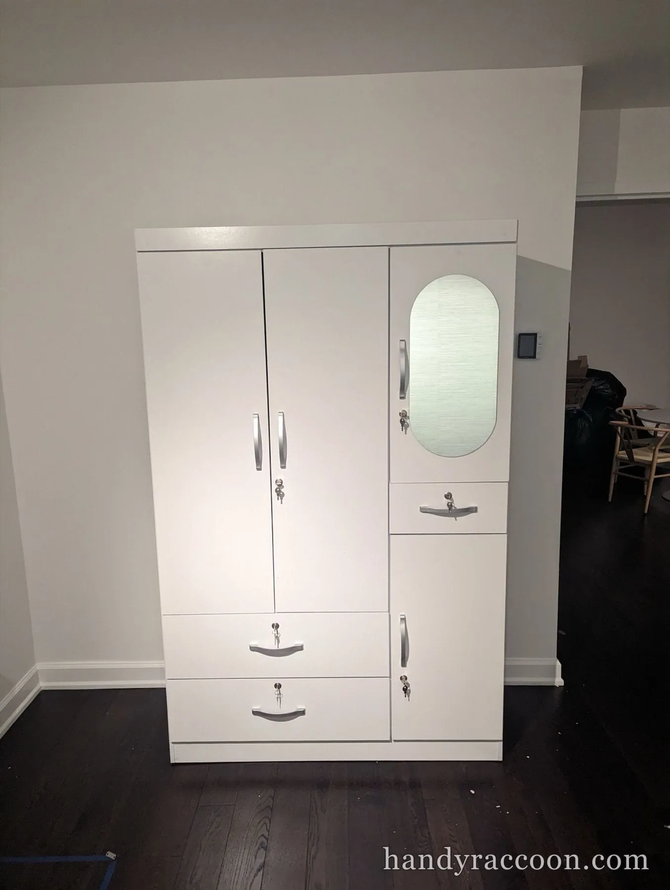 White three-door wardrobe with an oval mirror, drawers and keyed locks on a dark floor.