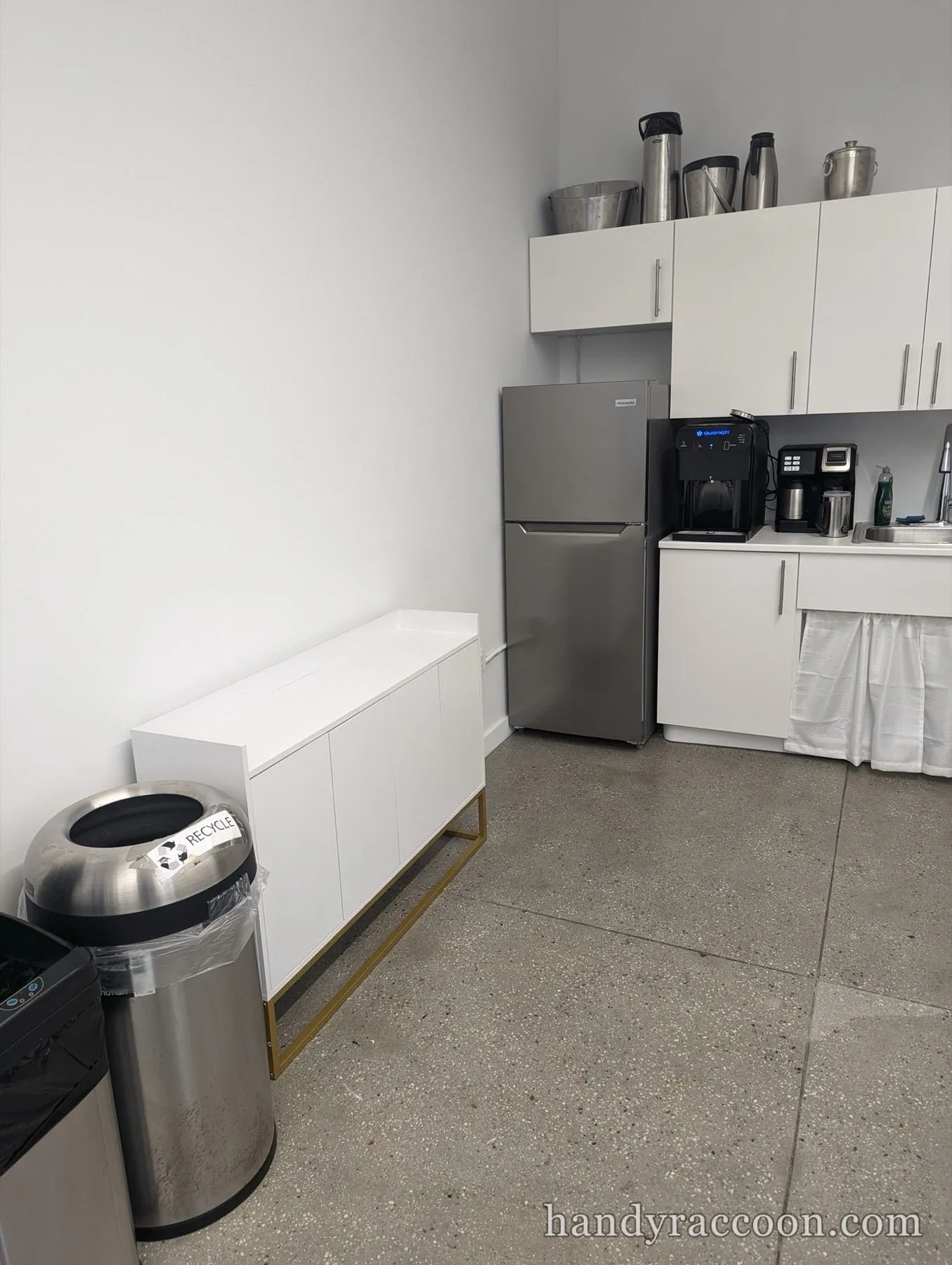 Low white cabinet on a gold metal frame against the wall of an office kitchenette with a fridge.