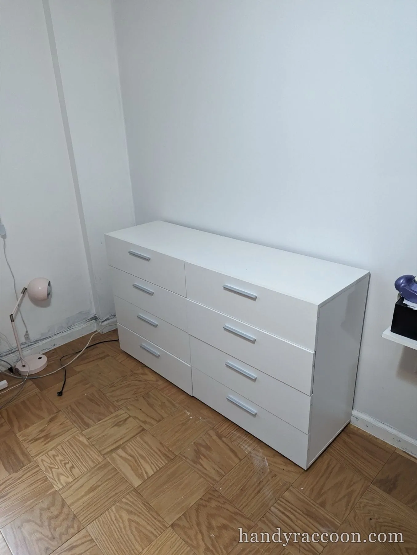 White eight-drawer double dresser standing against a white wall on a parquet floor.