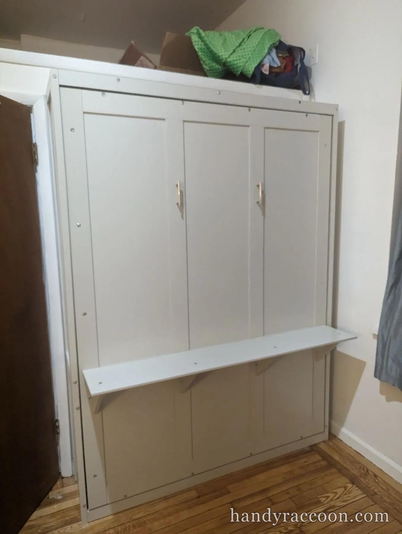 White vertical murphy wall bed cabinet with a folded-down shelf, in a corner on a wood floor.