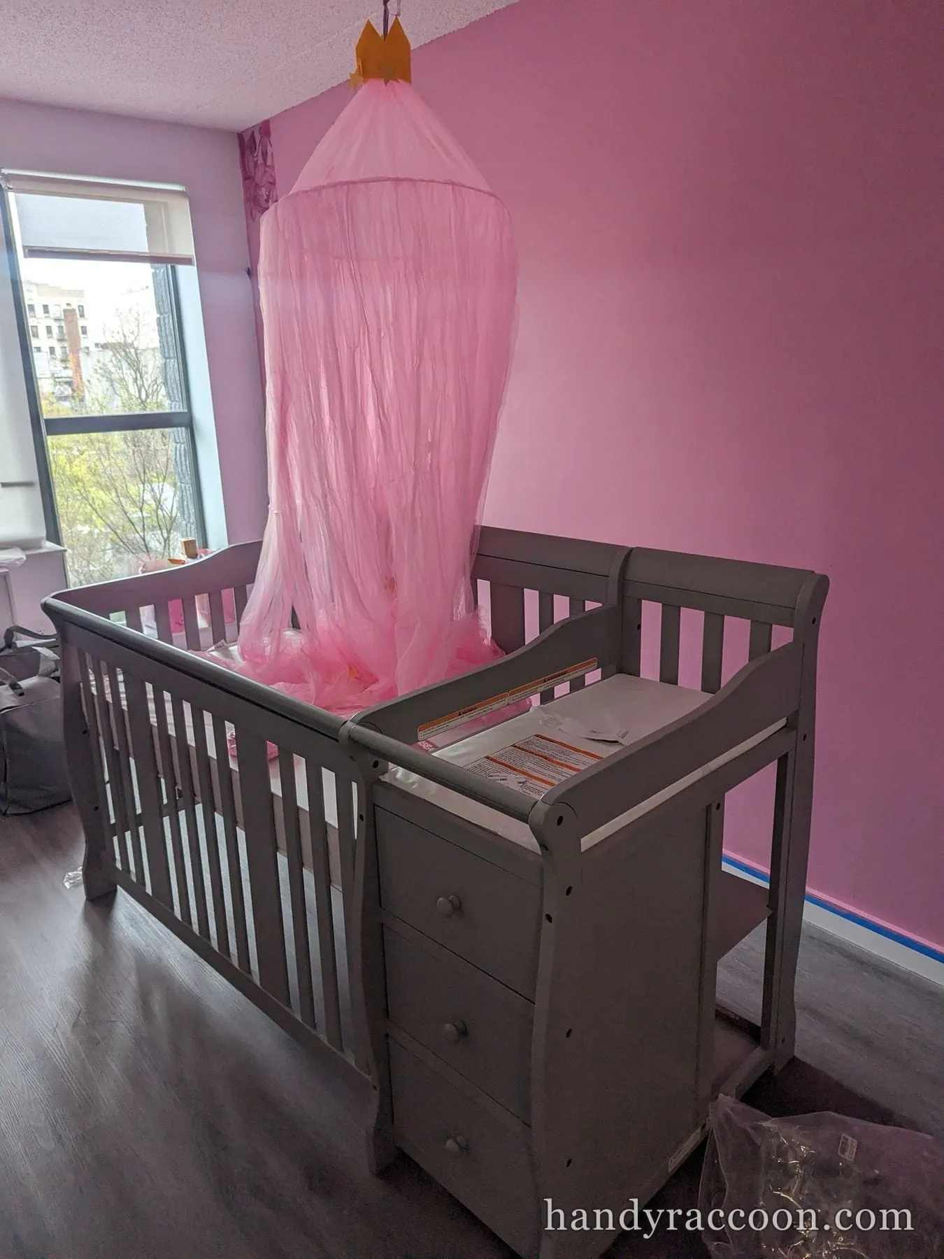 Gray crib with attached changing table and pink canopy assembled in a pink nursery by a window.