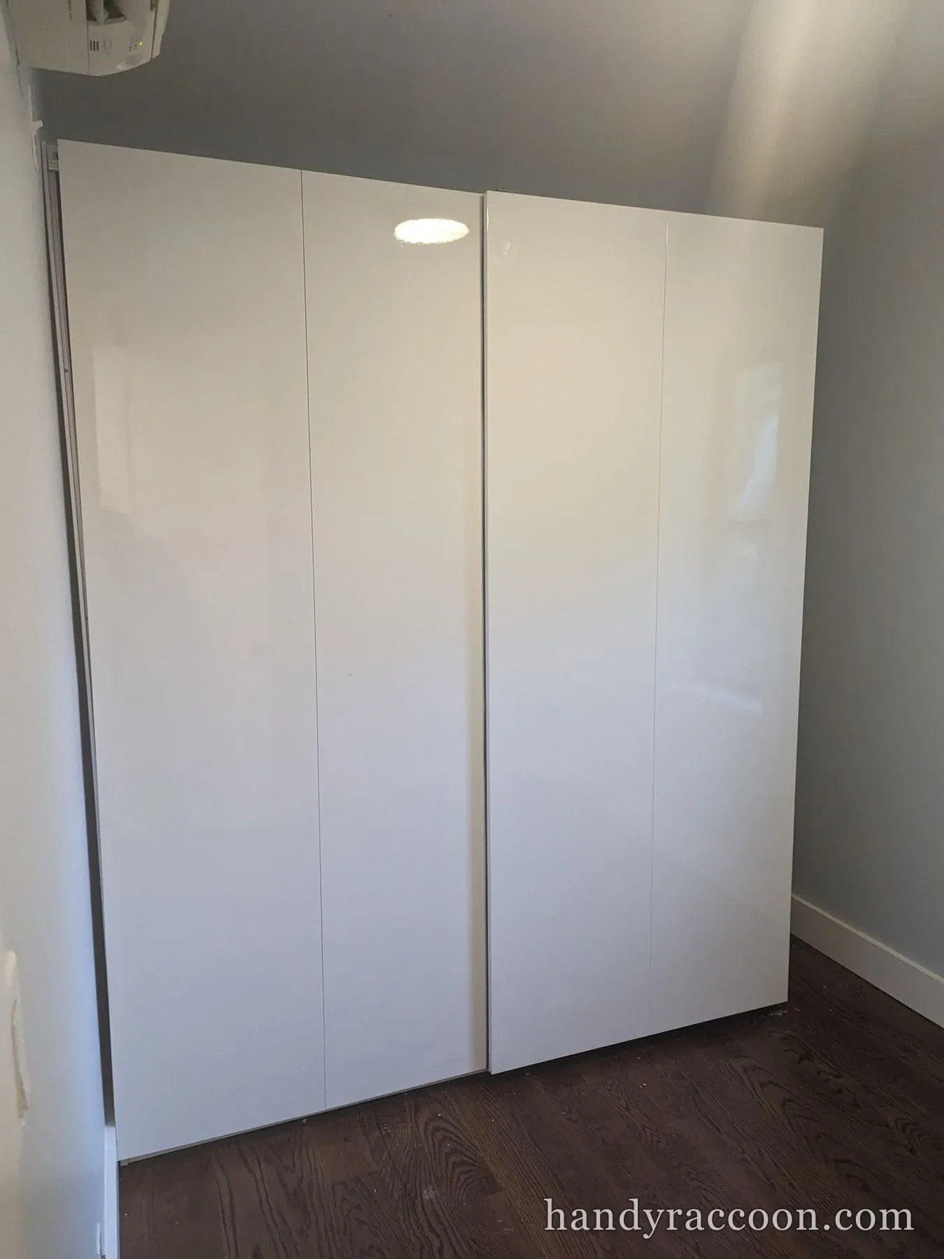 High-gloss white two-door wardrobe assembled and standing against a gray wall.