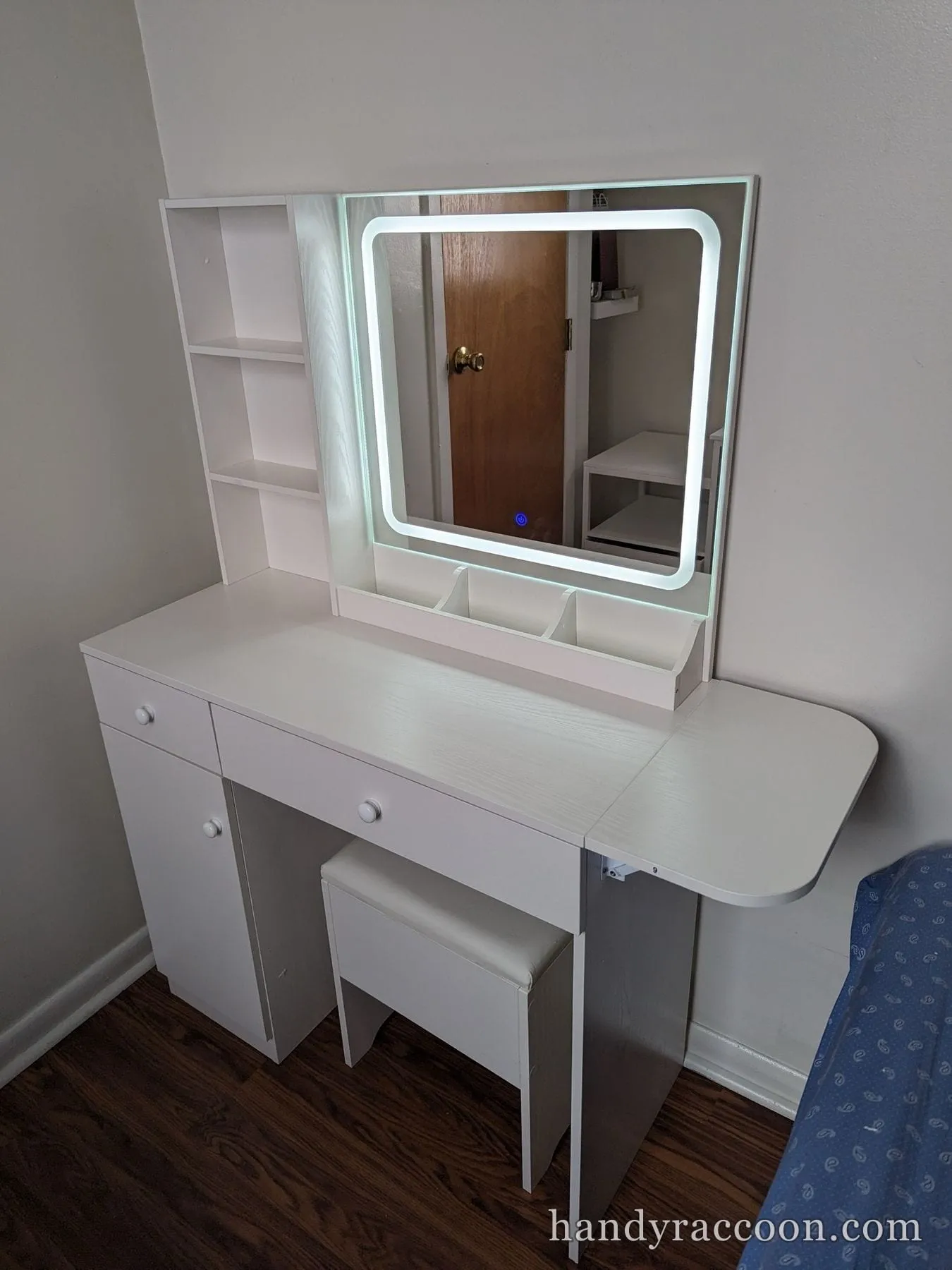 White vanity desk with a lit square mirror, shelving, drawers, and stool assembled.